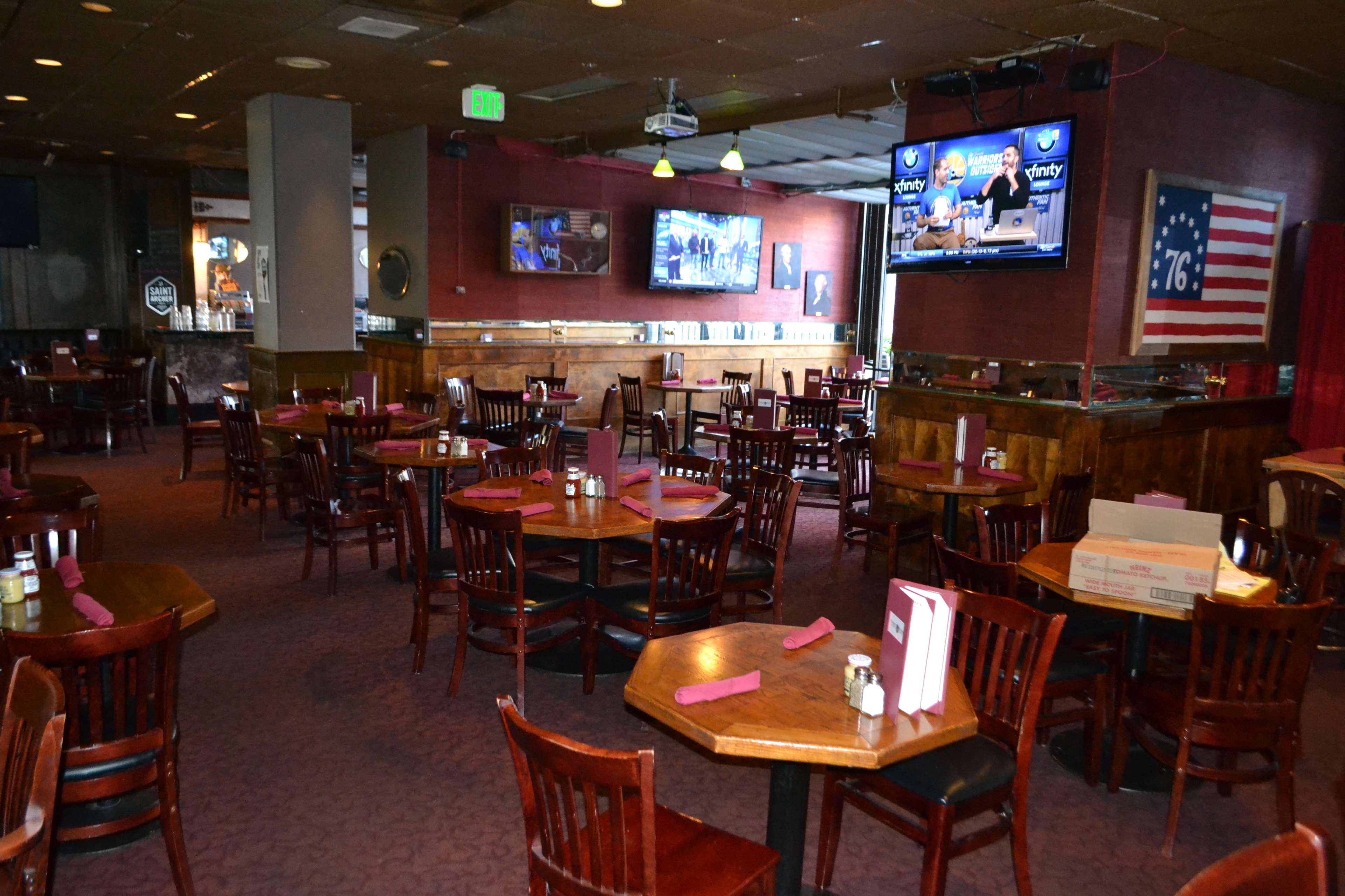 A mostly empty restaurant with wooden tables and chairs, two televisions mounted on the walls, and a large American flag displayed.