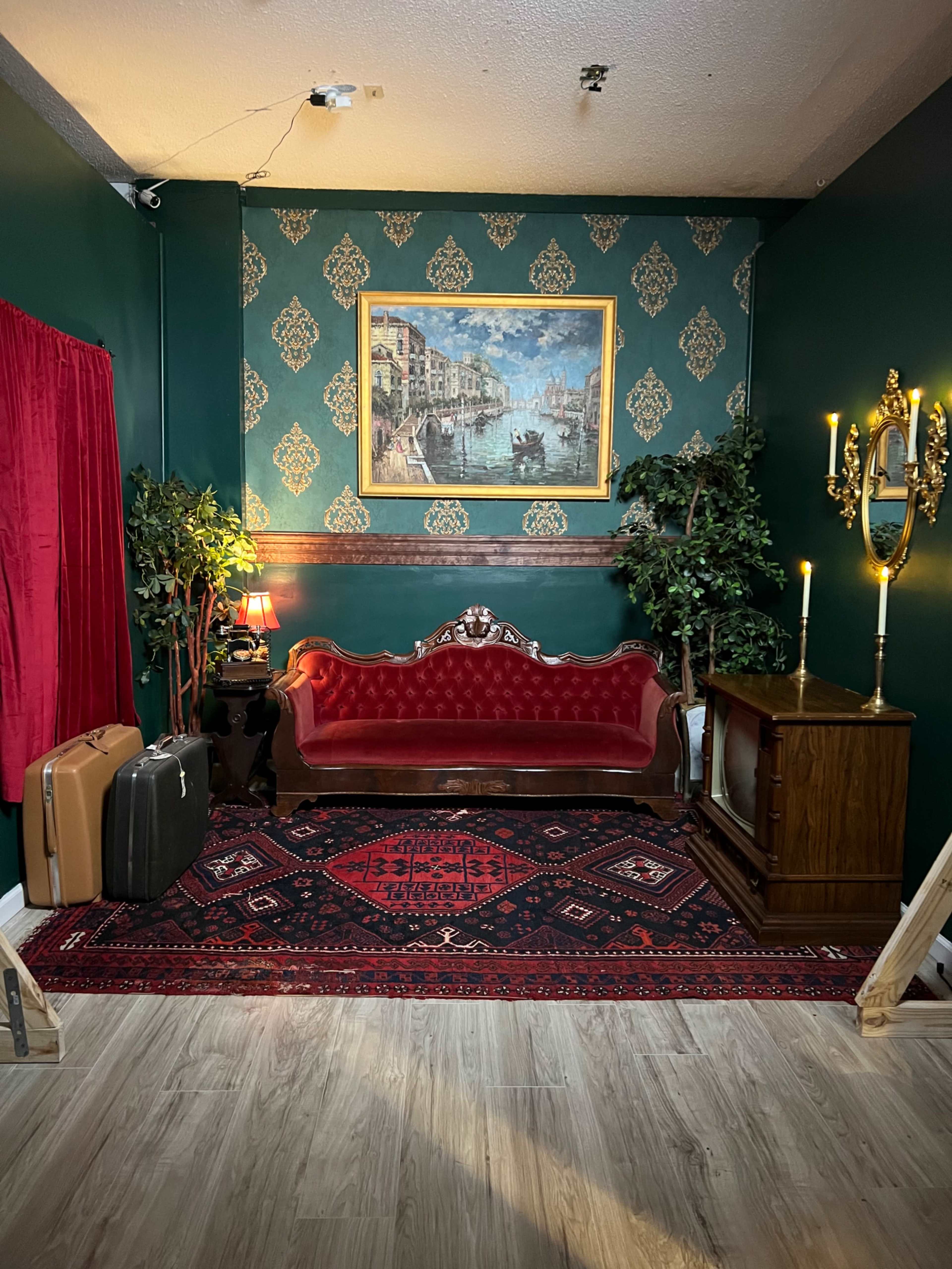 A vintage-inspired living room with a red velvet sofa, an ornate gold-framed painting, and a wooden television set against a patterned green wall.