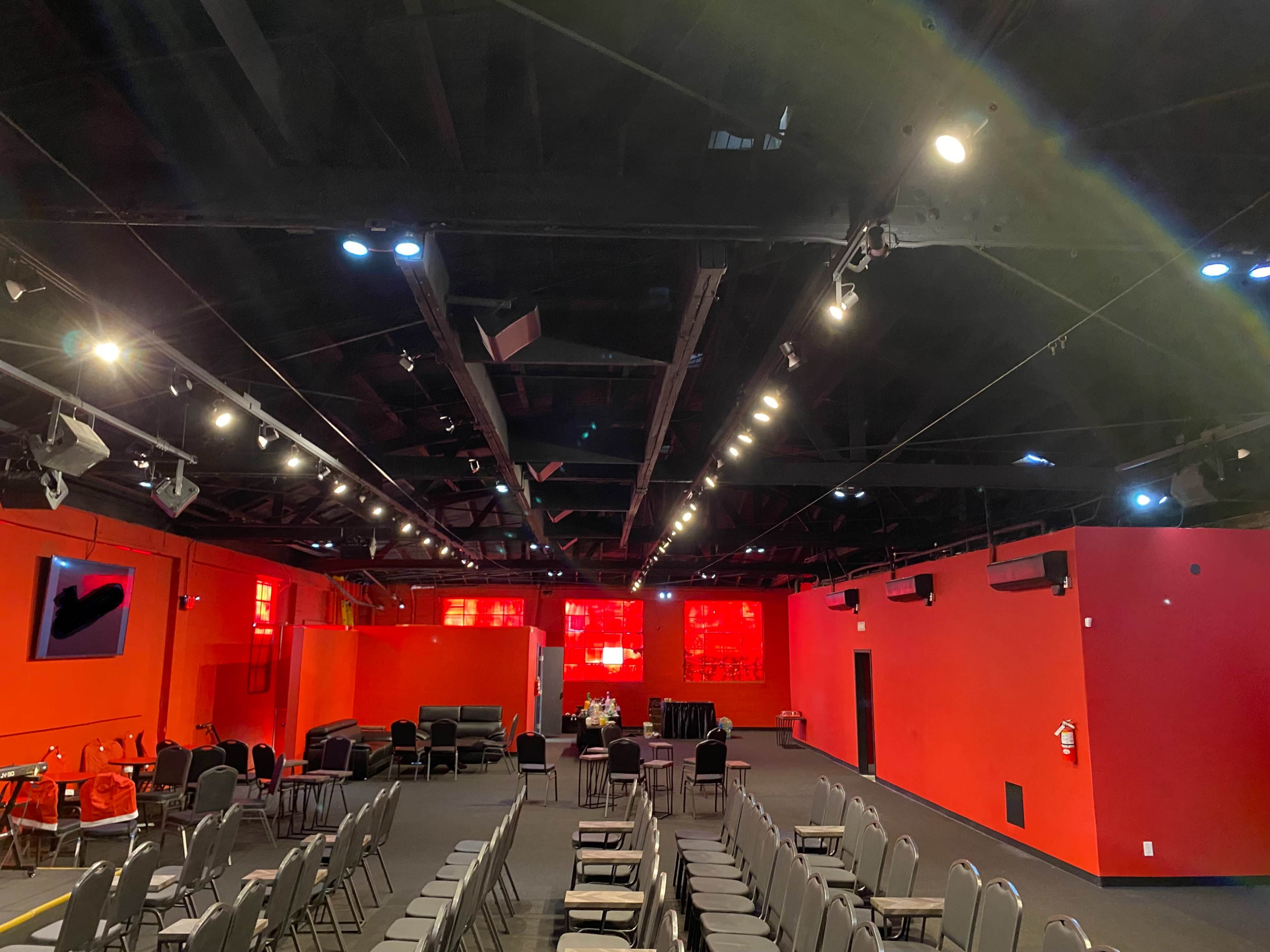 The image shows a large room with red walls, multiple rows of metal chairs arranged facing a stage, and bright lighting overhead.