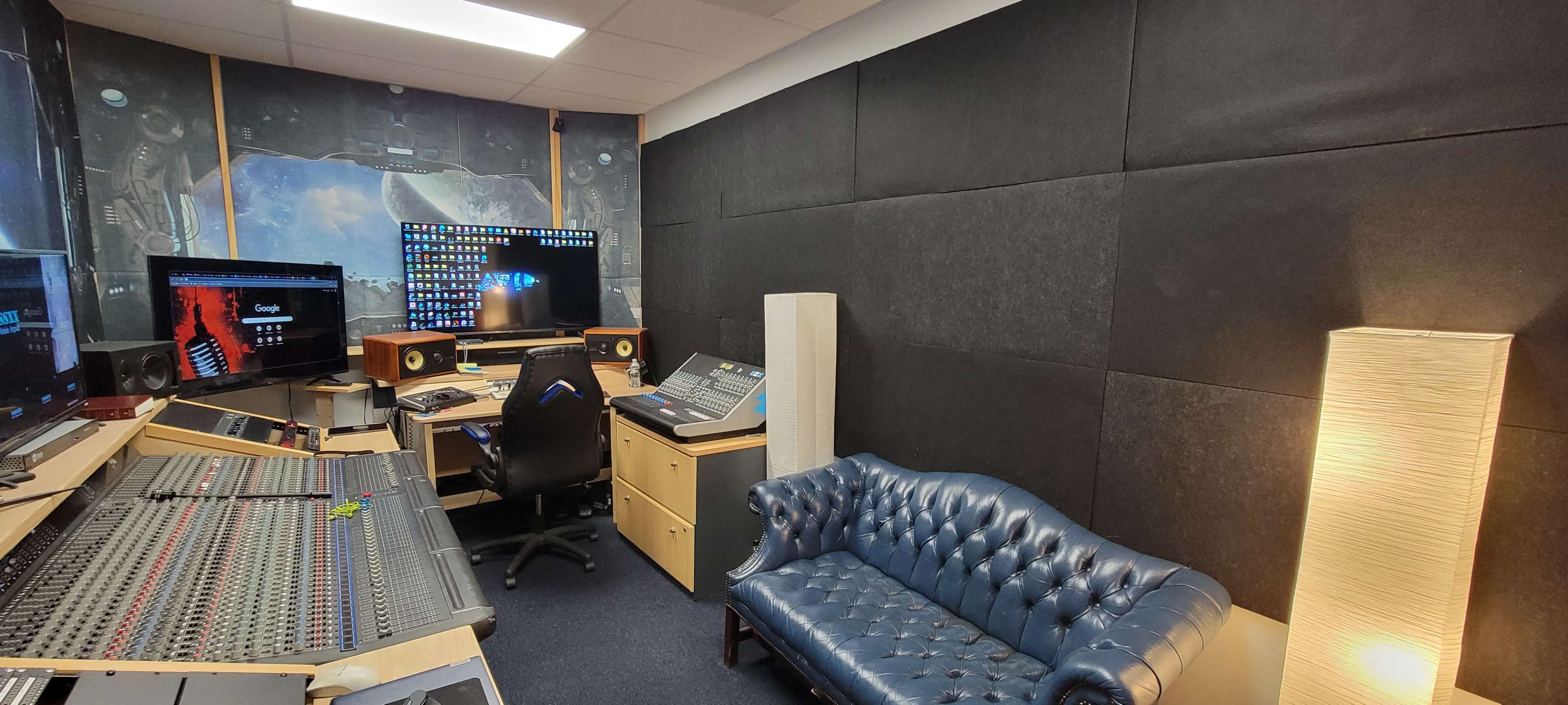 A sound studio featuring a control desk with various audio equipment, a computer monitor displaying a digital interface, a black executive chair, a blue leather couch, and acoustic panels on the walls.