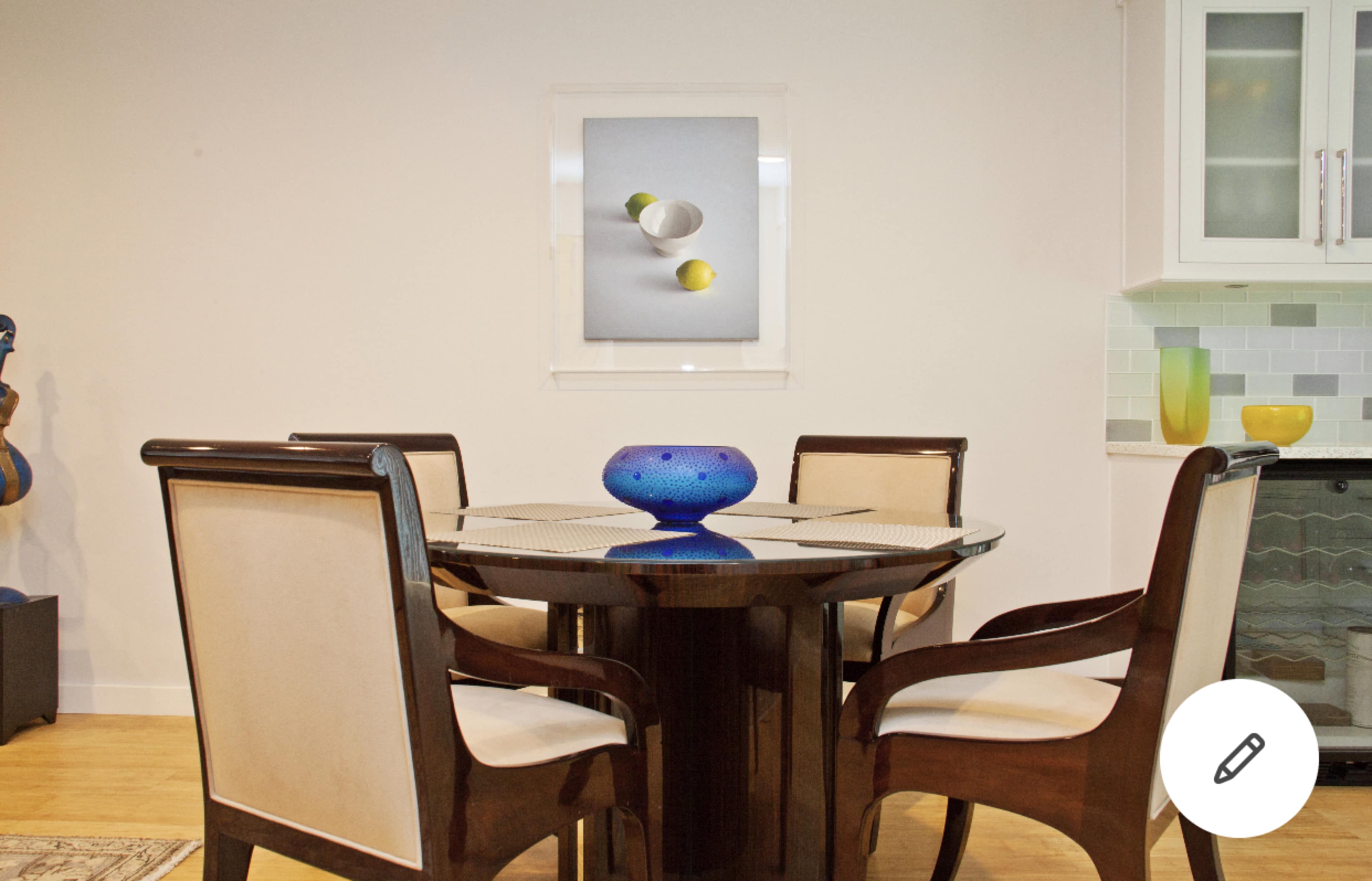 A round wooden dining table is set with four upholstered chairs, featuring a blue decorative bowl at the center and a wall-mounted art piece above.