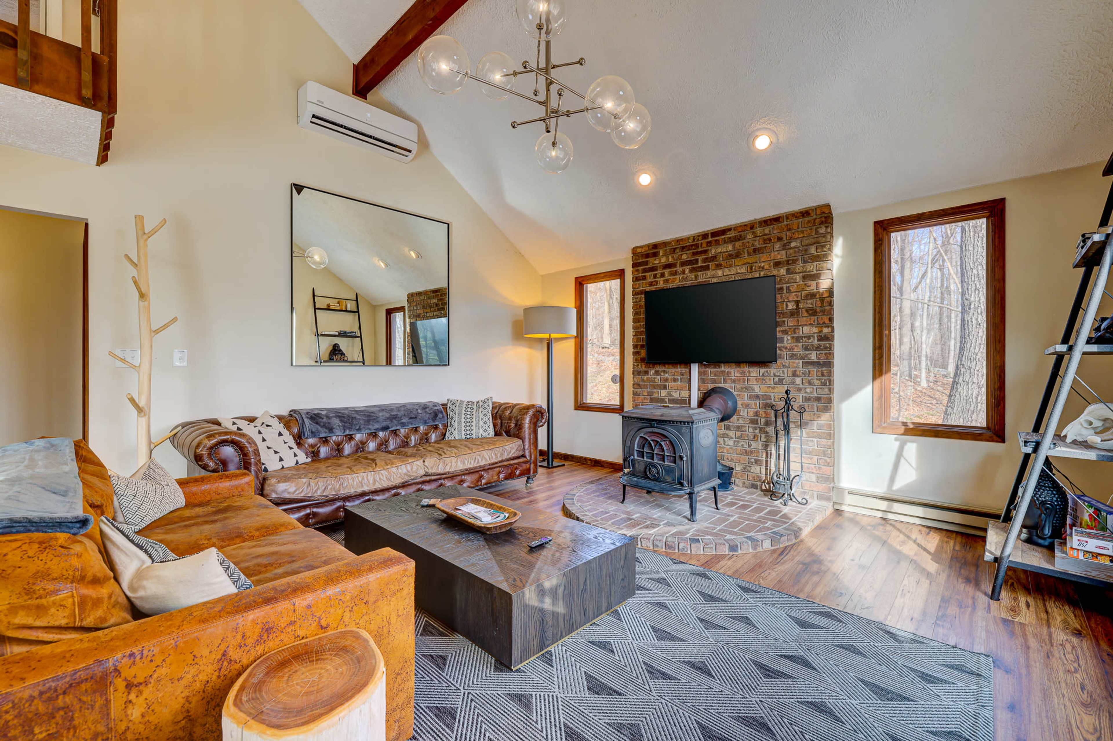 A modern living room features a leather sofa, a wooden coffee table, a brick fireplace, and large windows overlooking a wooded area.
