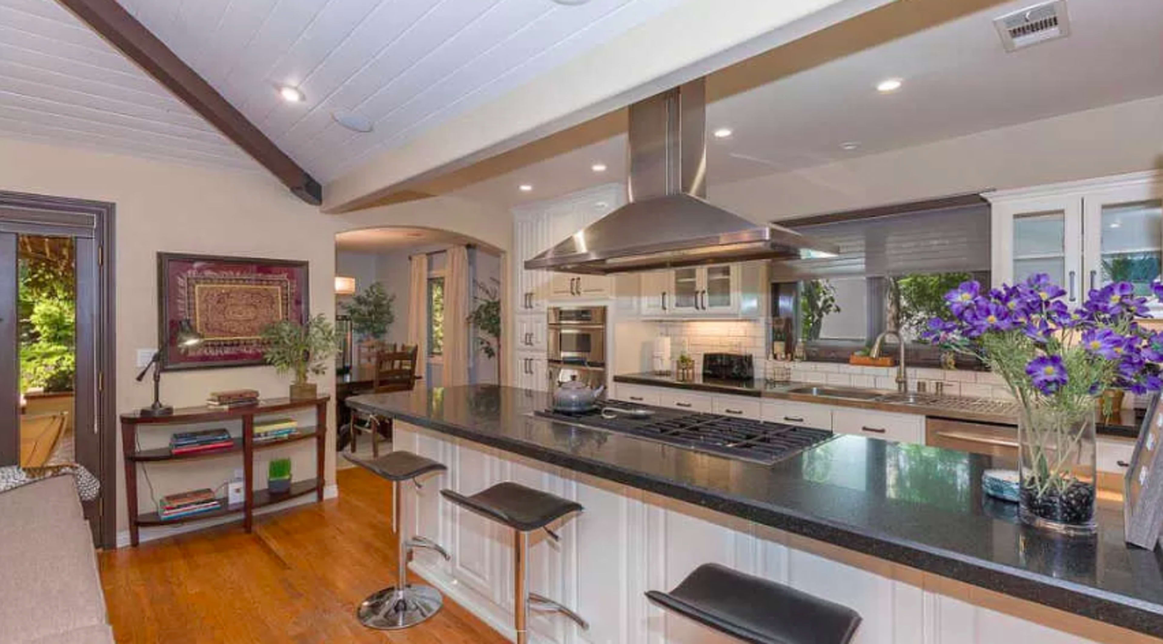 The image shows a modern kitchen with a large island, stainless steel appliances, and bar stools, featuring wooden flooring and natural light.