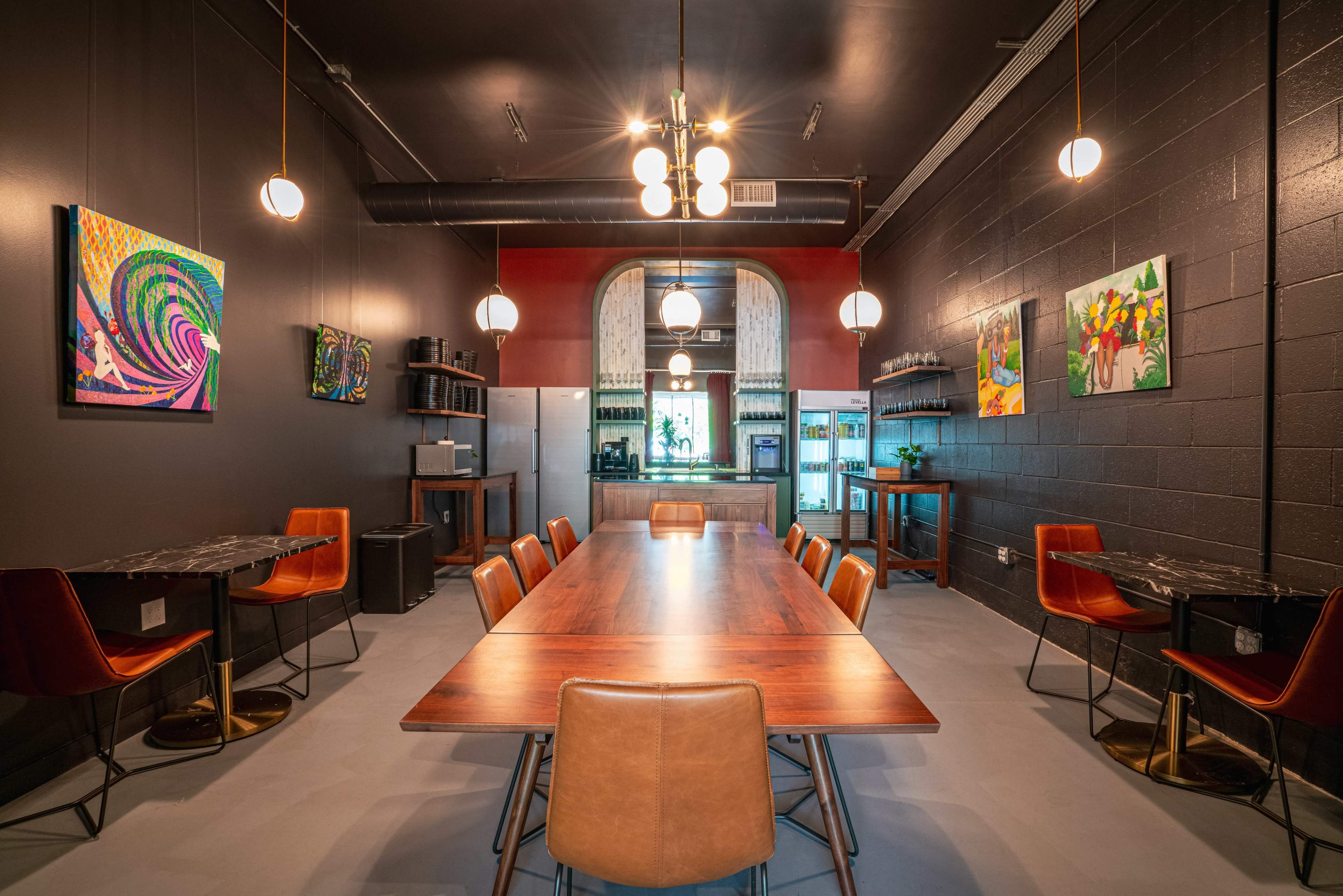 A modern dining or meeting space with a long wooden table surrounded by chairs, illuminated by pendant lights, and adorned with colorful artwork on the black walls.
