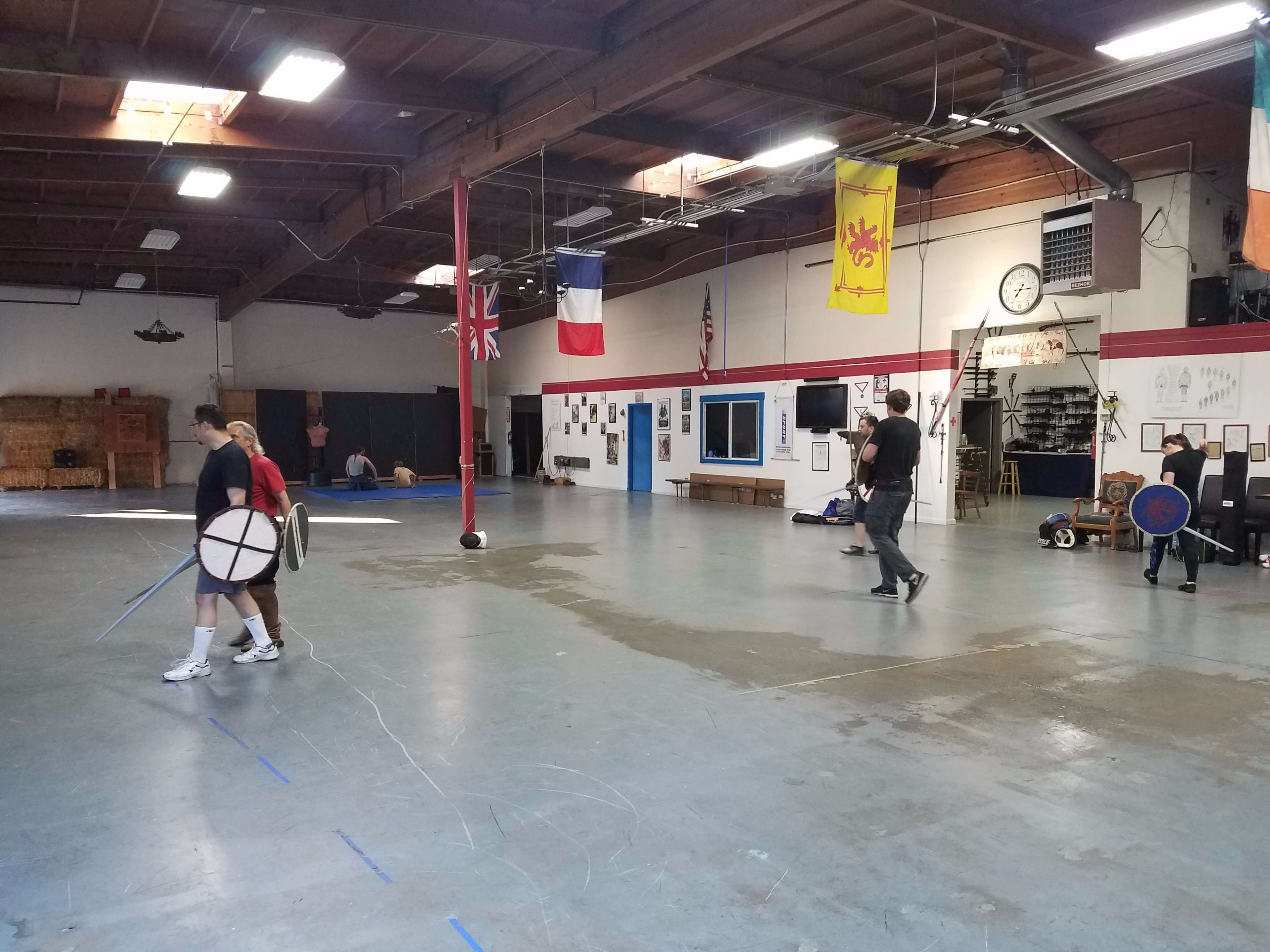 A spacious indoor training facility features various wall displays and flags, while individuals practice swordsmanship and other skills across the floor.