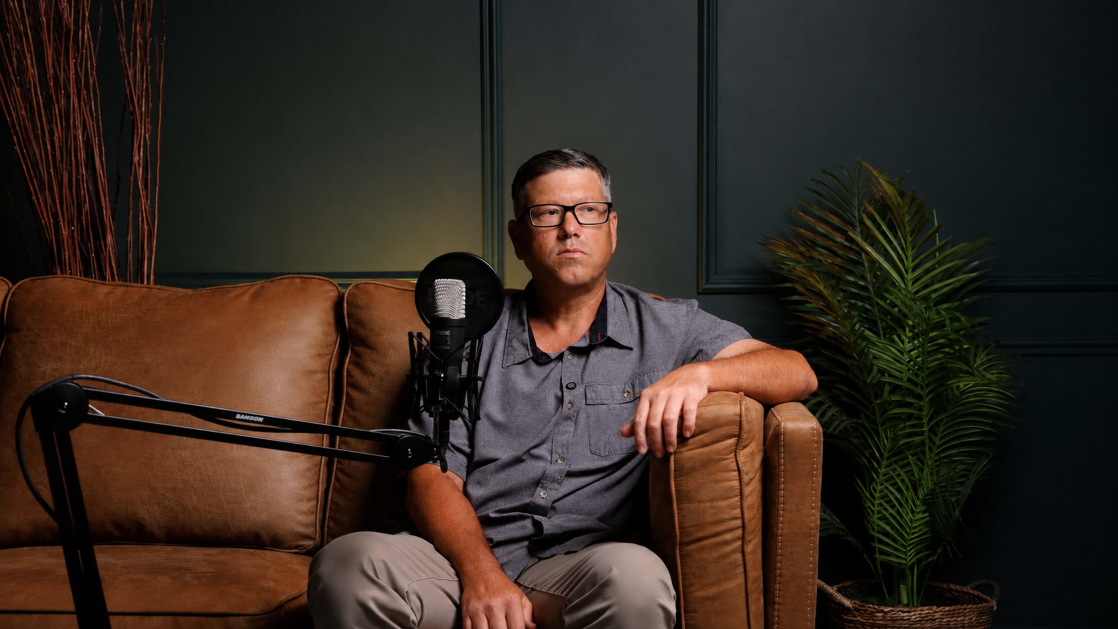 A man wearing glasses is sitting on a brown leather couch with a microphone mounted on an arm beside him.