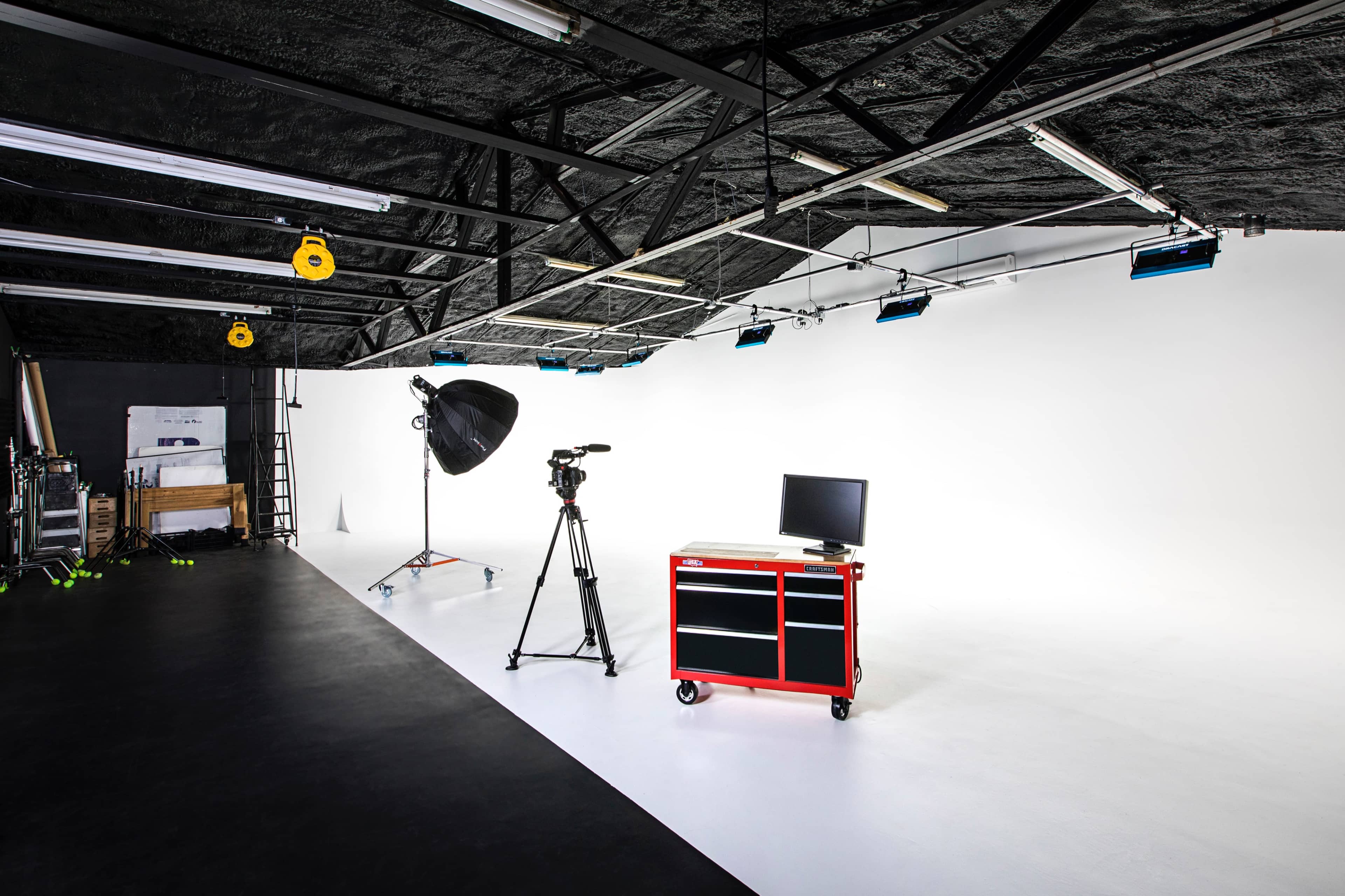 A photography studio with a camera on a tripod, a large light umbrella, a red tool cart, and a monitor against a white backdrop.