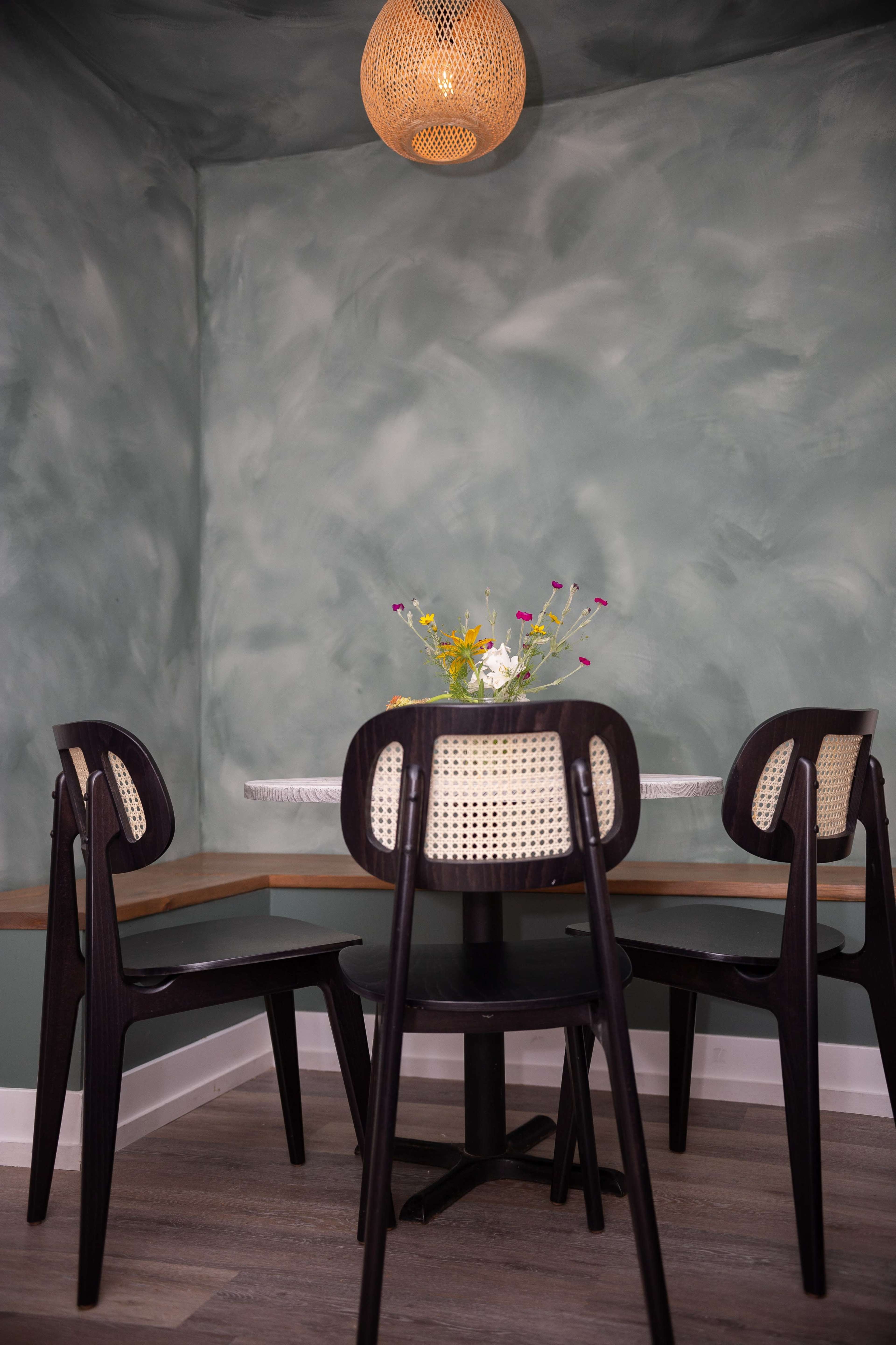 A small dining area features a round table surrounded by three black chairs with woven backs, set against a textured green wall and illuminated by a pendant light.