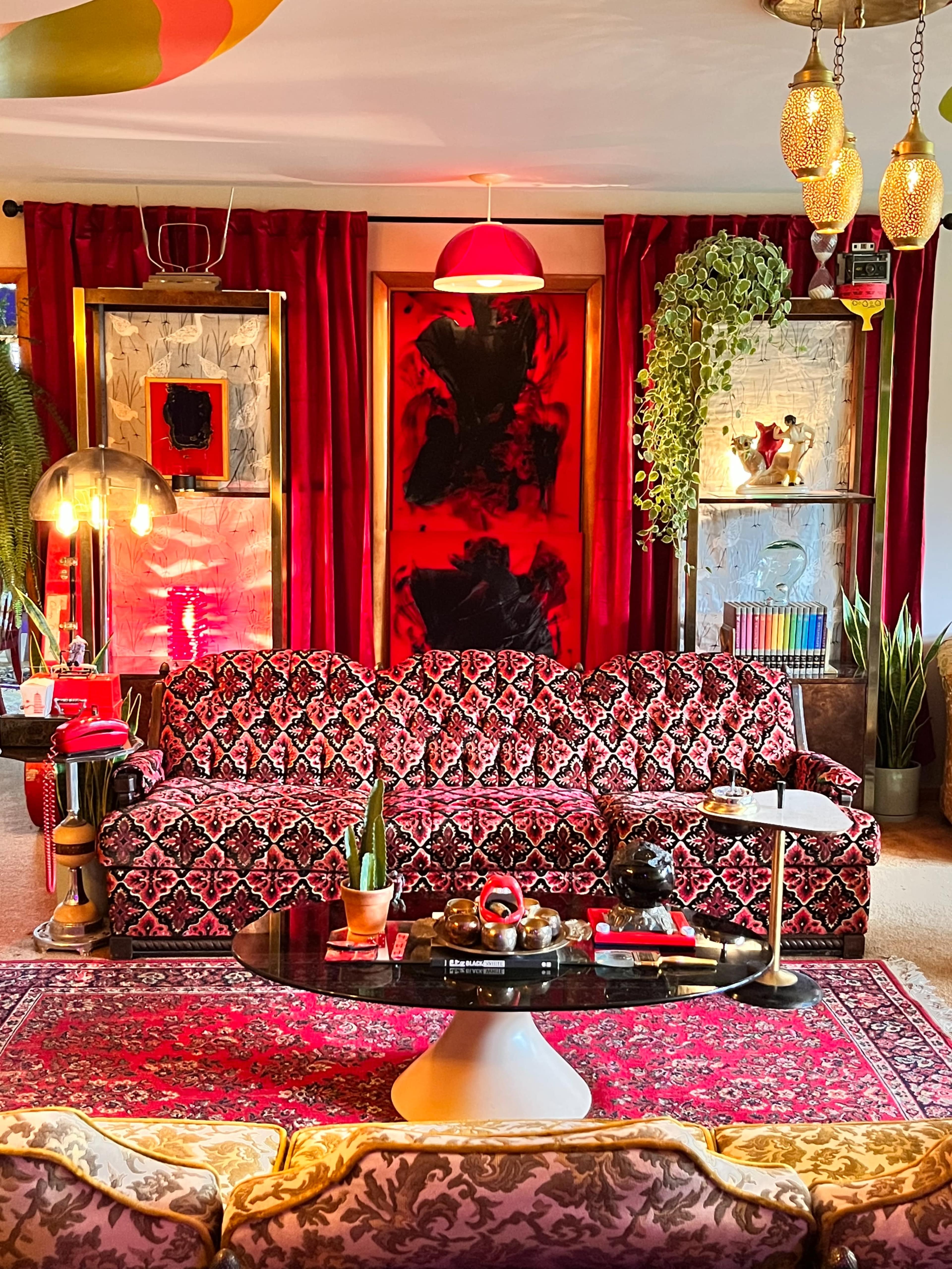 The image depicts a vibrant living room featuring a dark floral-patterned sofa, a round coffee table, elaborate red curtains, and various decorative plants and artwork.