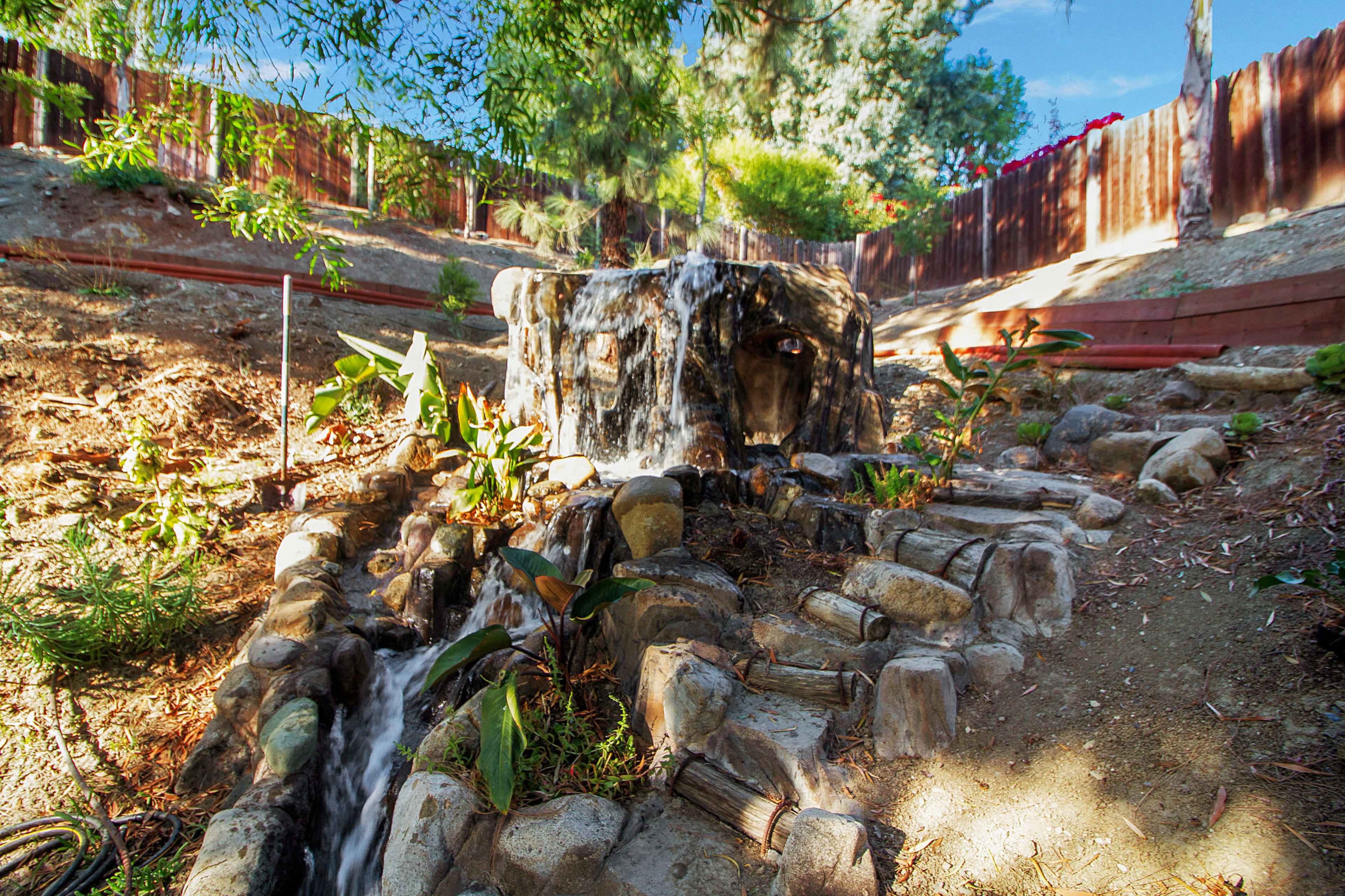 A stone fountain flows water down a landscaped slope surrounded by various plants and trees.