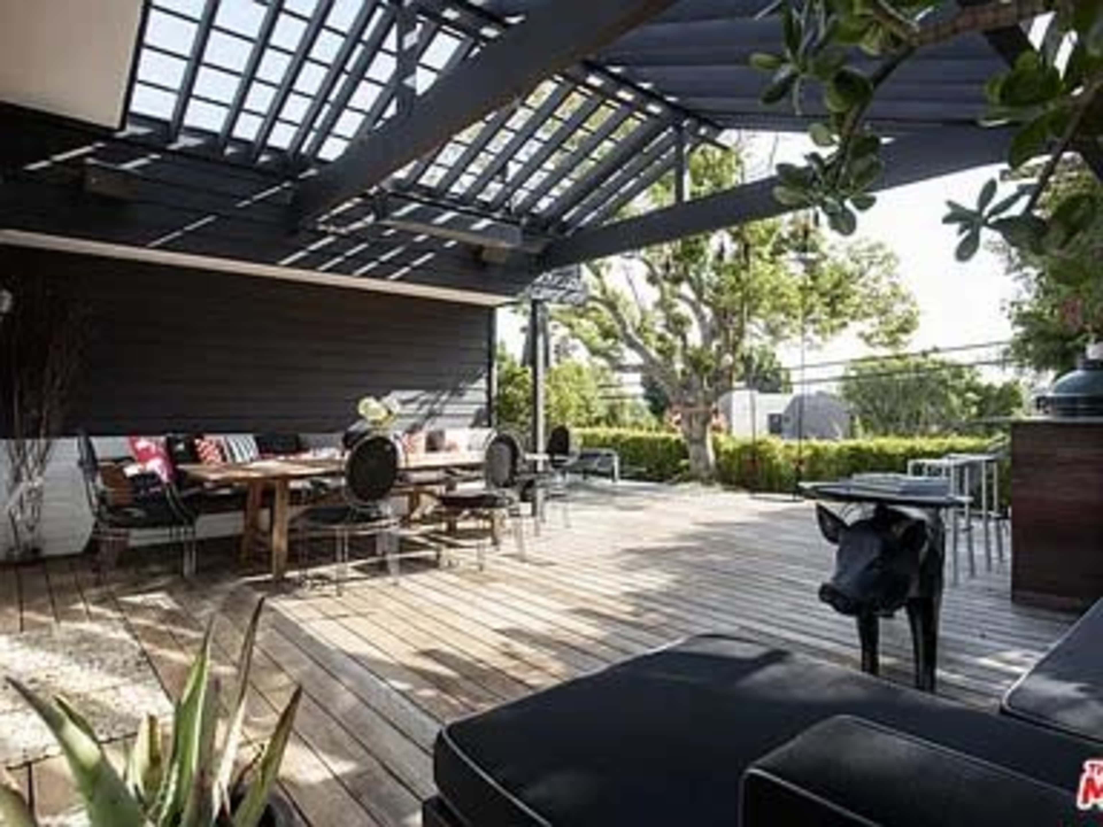 A wooden-decked outdoor living space with a dining area, seating, and a trellis overhead, surrounded by greenery.