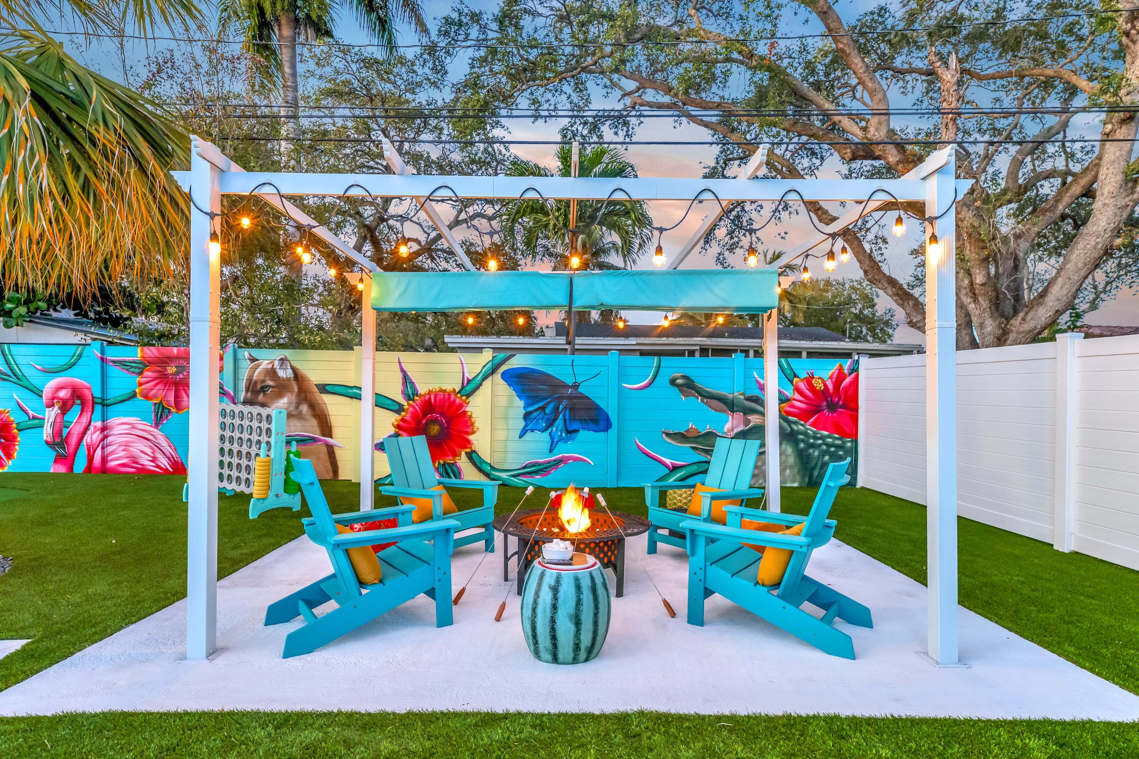 The image shows a colorful outdoor seating area with blue chairs arranged around a fire pit, surrounded by vibrant mural art and decorative string lights.