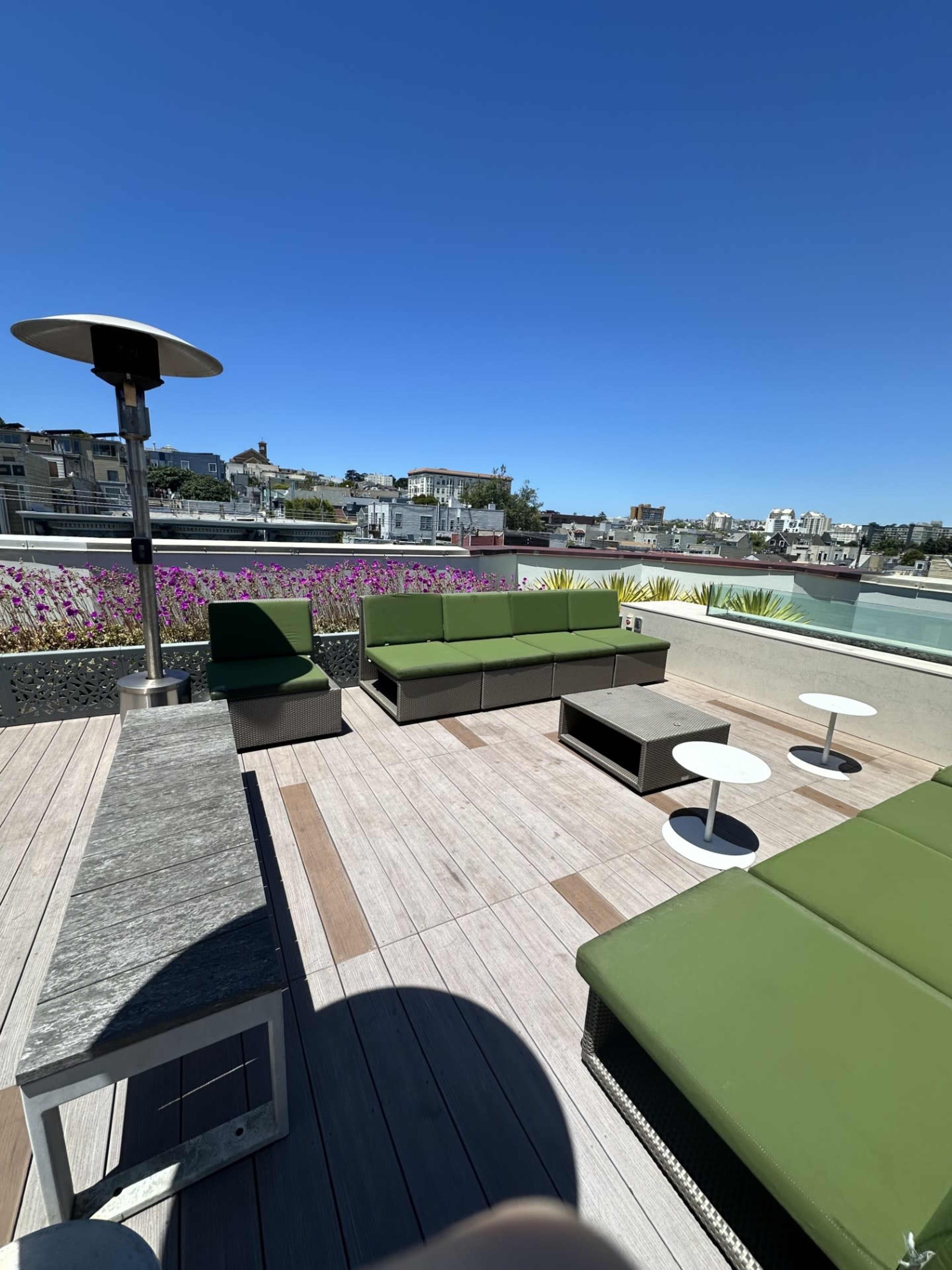 A rooftop terrace with green lounge furniture, wooden decking, and a view of buildings in the background under a clear blue sky.