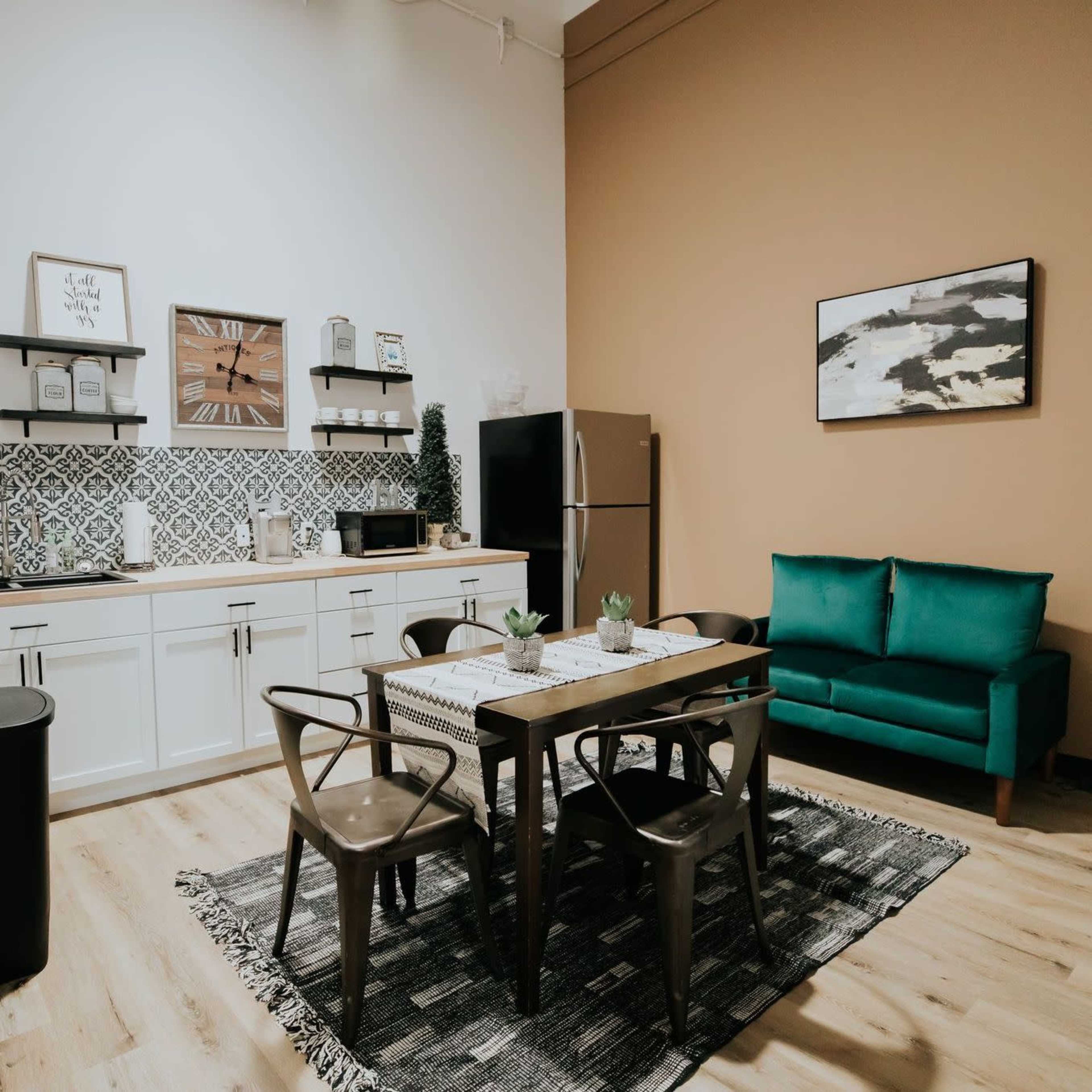 A modern kitchen and dining area features a metal dining table with chairs, a green sofa, and a patterned rug against a tan wall.