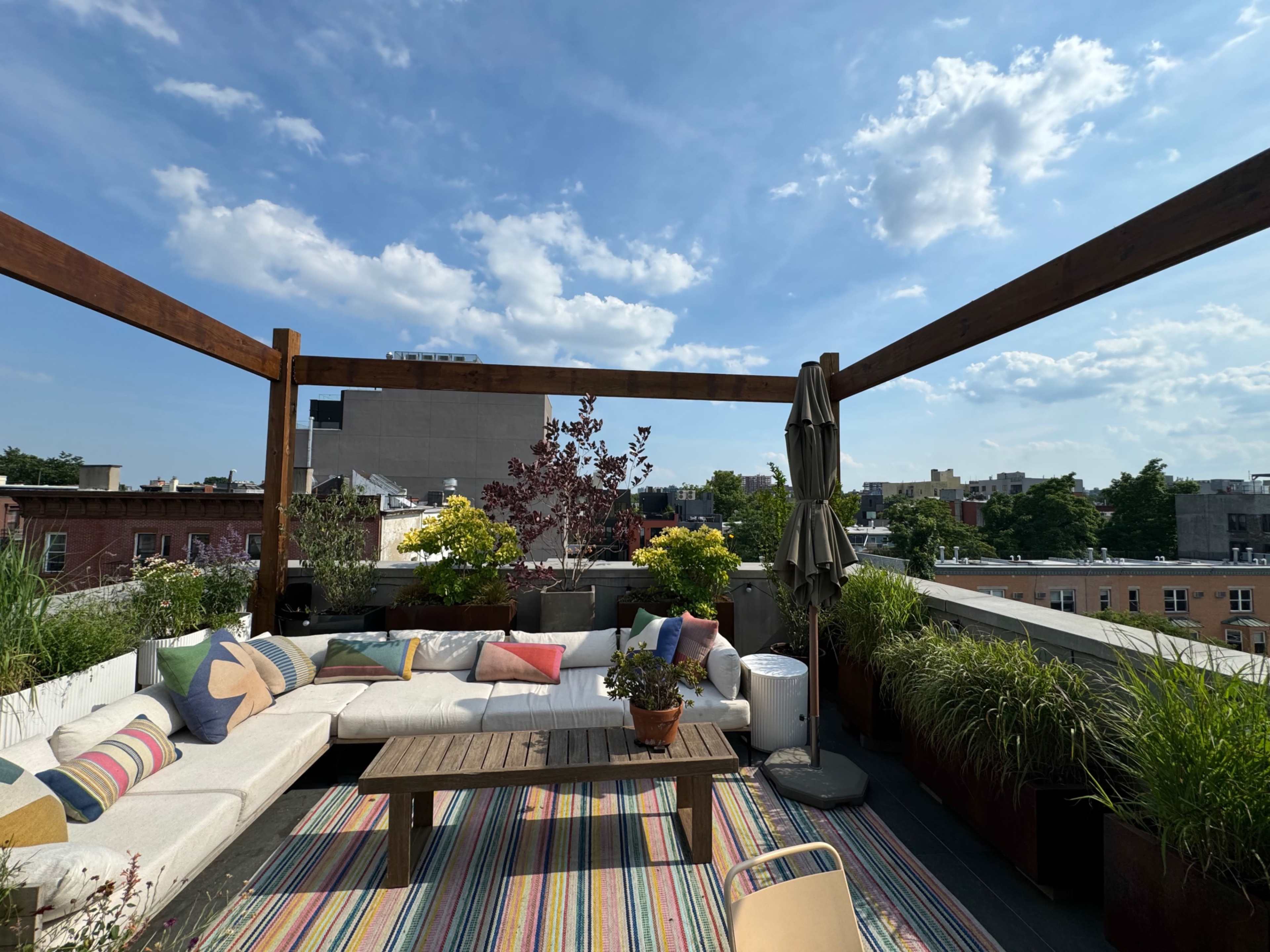 A rooftop terrace features a sectional couch with colorful cushions, a wooden coffee table, and potted plants under a partly cloudy sky.