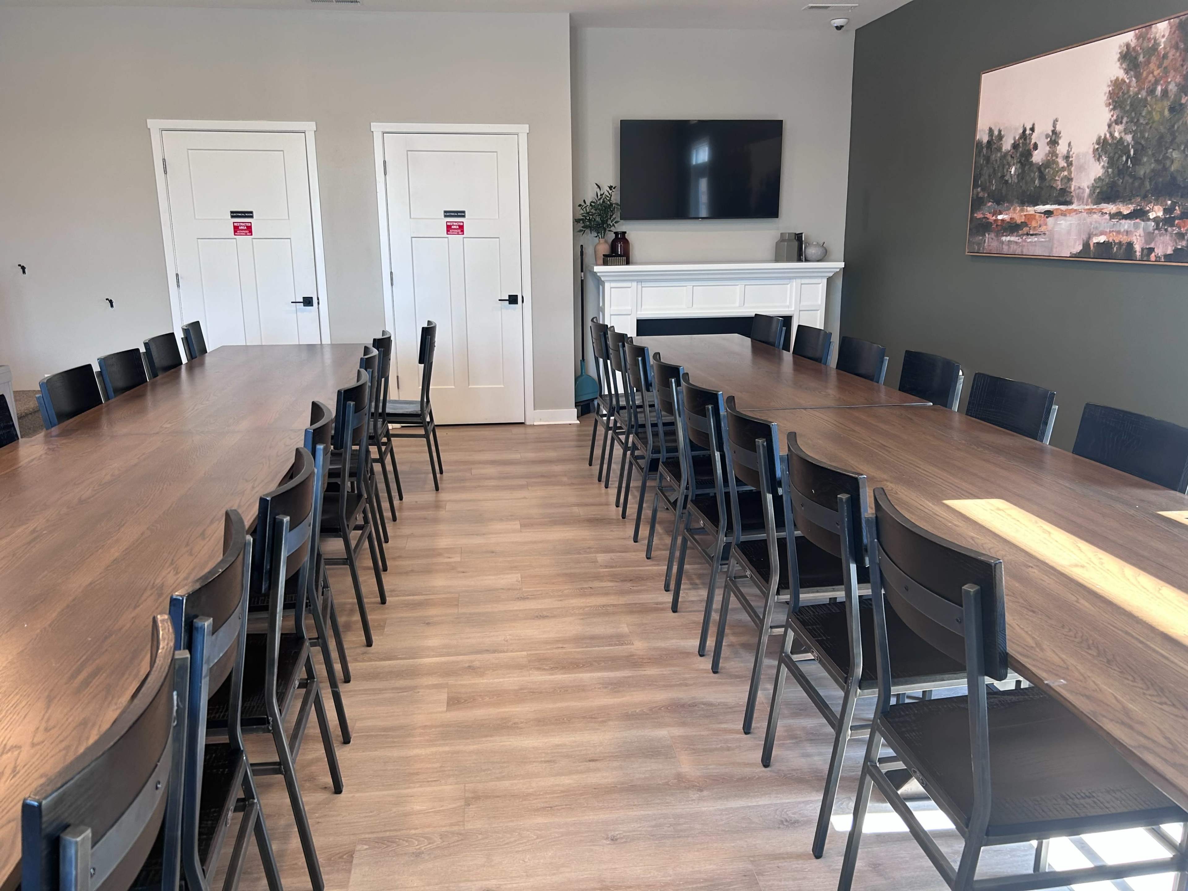 The image shows a long rectangular table surrounded by chairs in a meeting room with two doors and a large television on the wall.