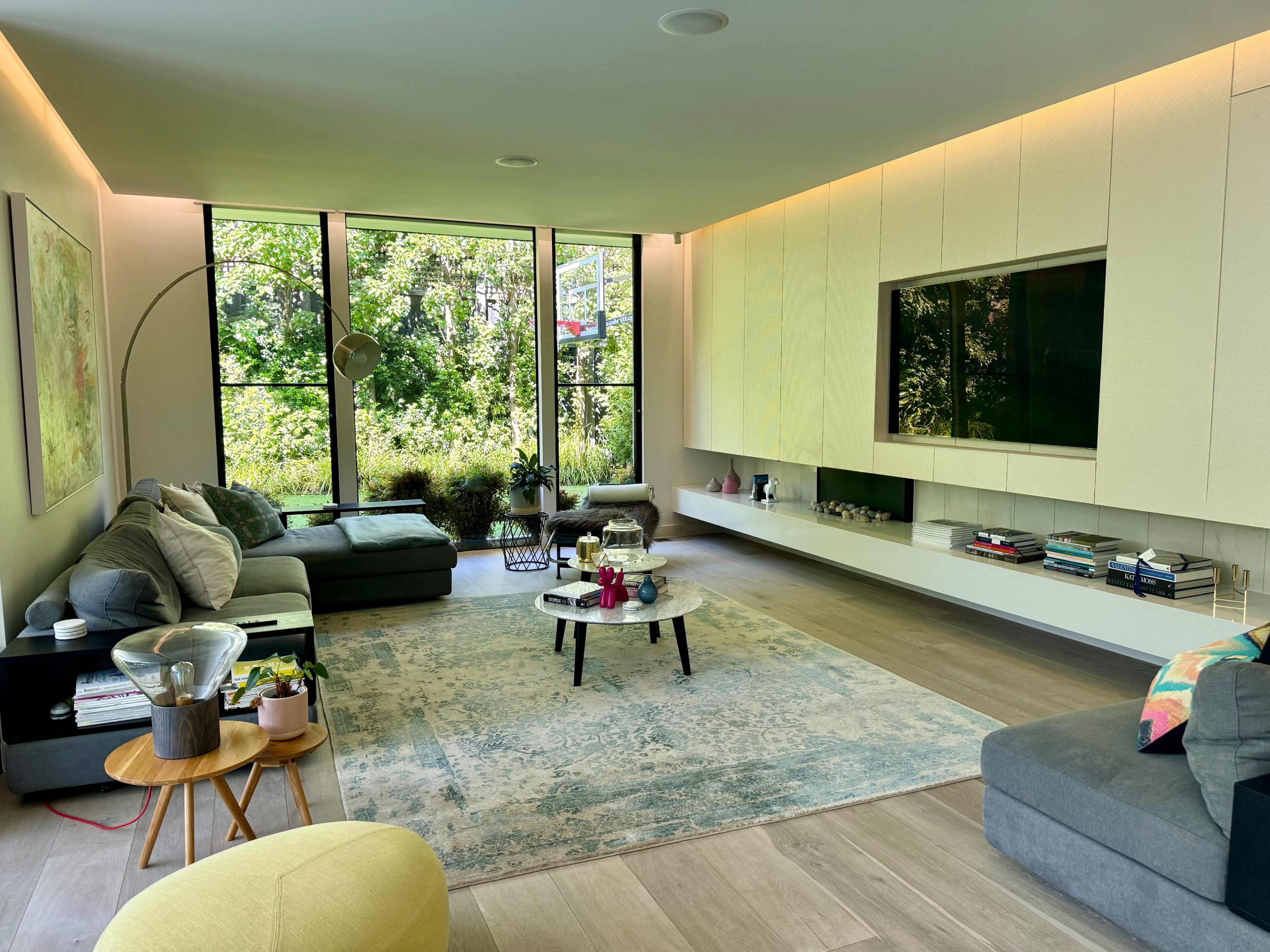 A modern living room with large windows overlooking greenery, featuring a sectional sofa, a round coffee table, and a wall-mounted TV.