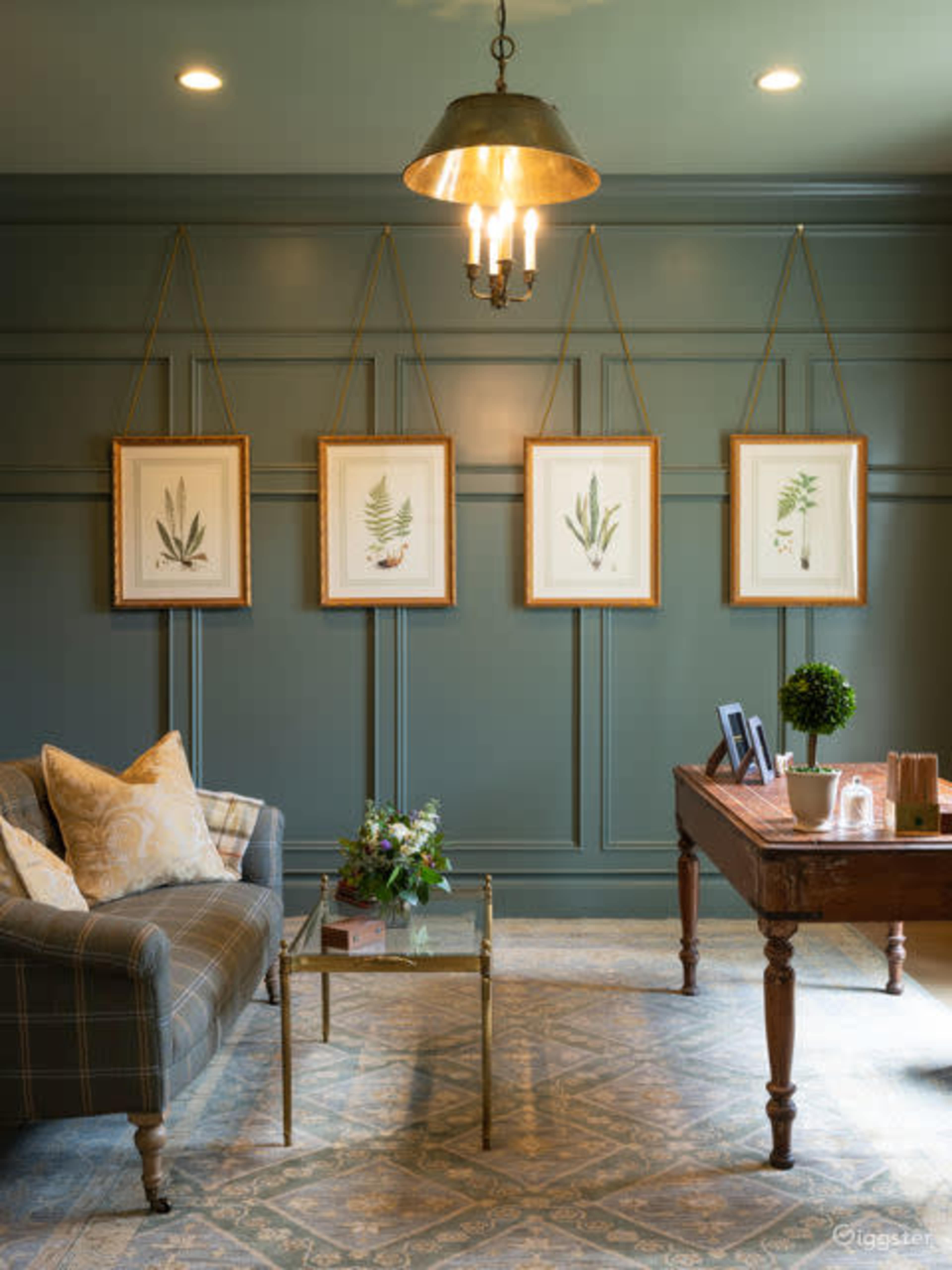 A cozy sitting area features a plaid sofa, a small table with a floral arrangement, and framed botanical prints hung on a dark green wall.