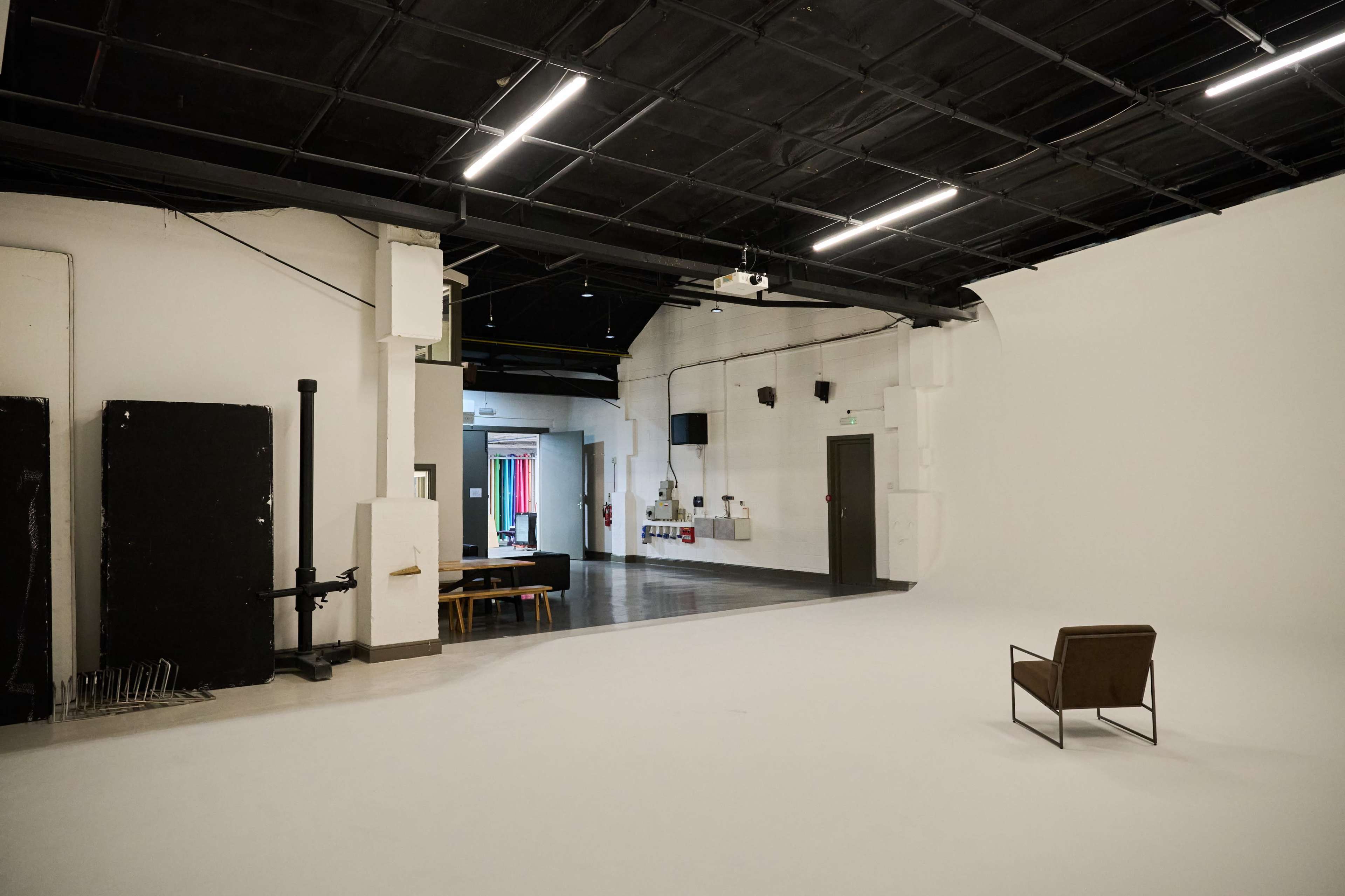 The image shows a spacious, minimalist studio with a single chair positioned on a white floor, surrounded by contrasting dark walls and empty spaces.