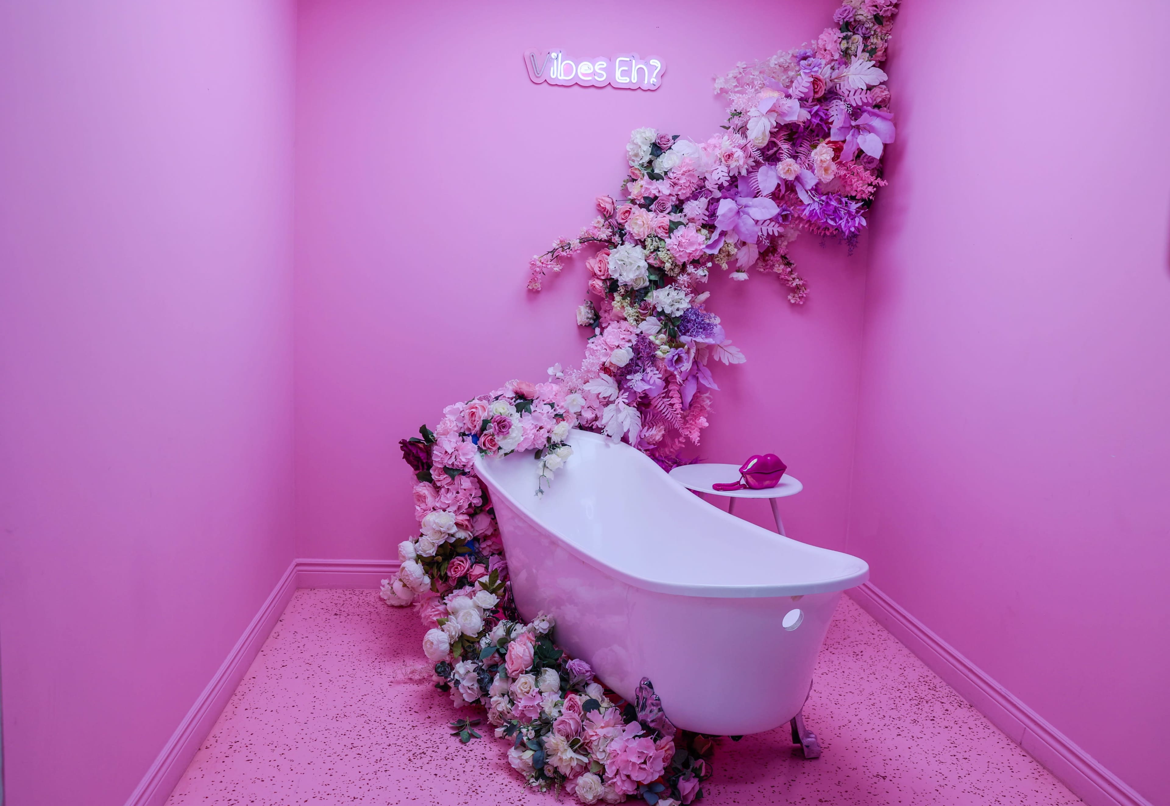 A white bathtub is surrounded by a cascading arrangement of artificial flowers against a bright pink wall, with a small table and a decorative sign reading "Vibes Eh?" above.