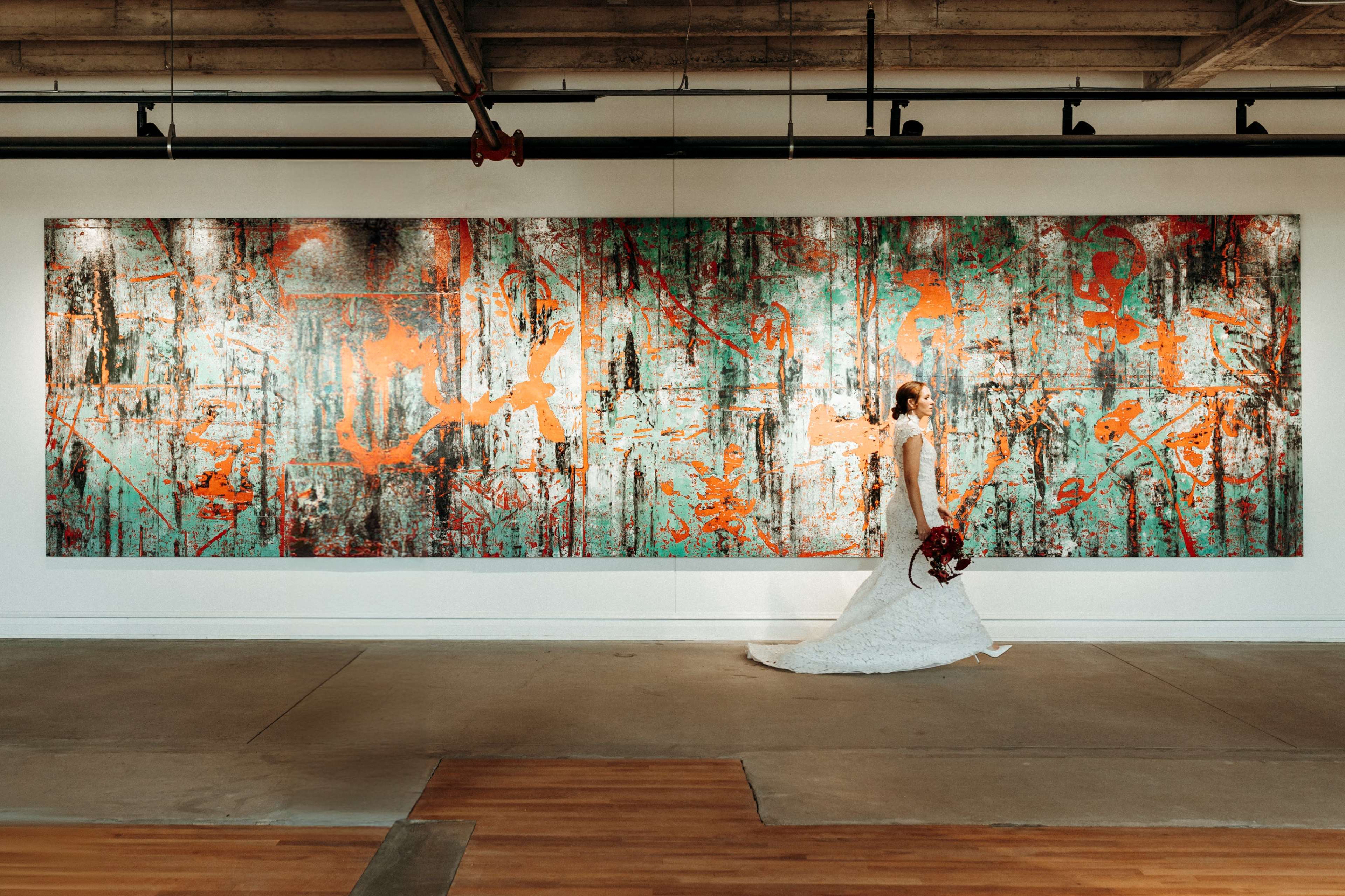 A bride in a wedding gown walks past a large, abstract painting with vibrant colors on a gallery wall.
