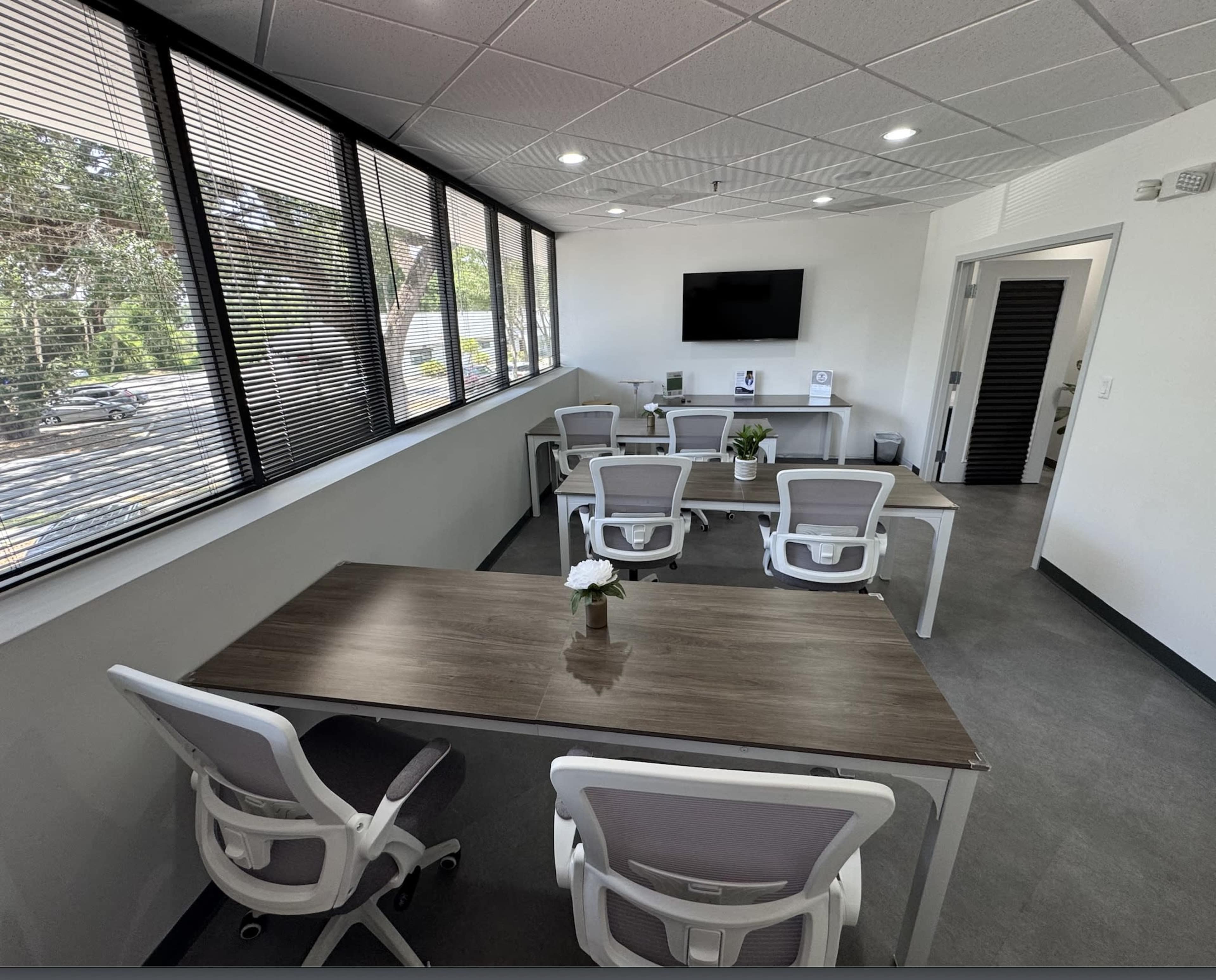 The image shows a modern office space featuring a large table with several white ergonomic chairs, large windows allowing natural light, and a wall-mounted television.