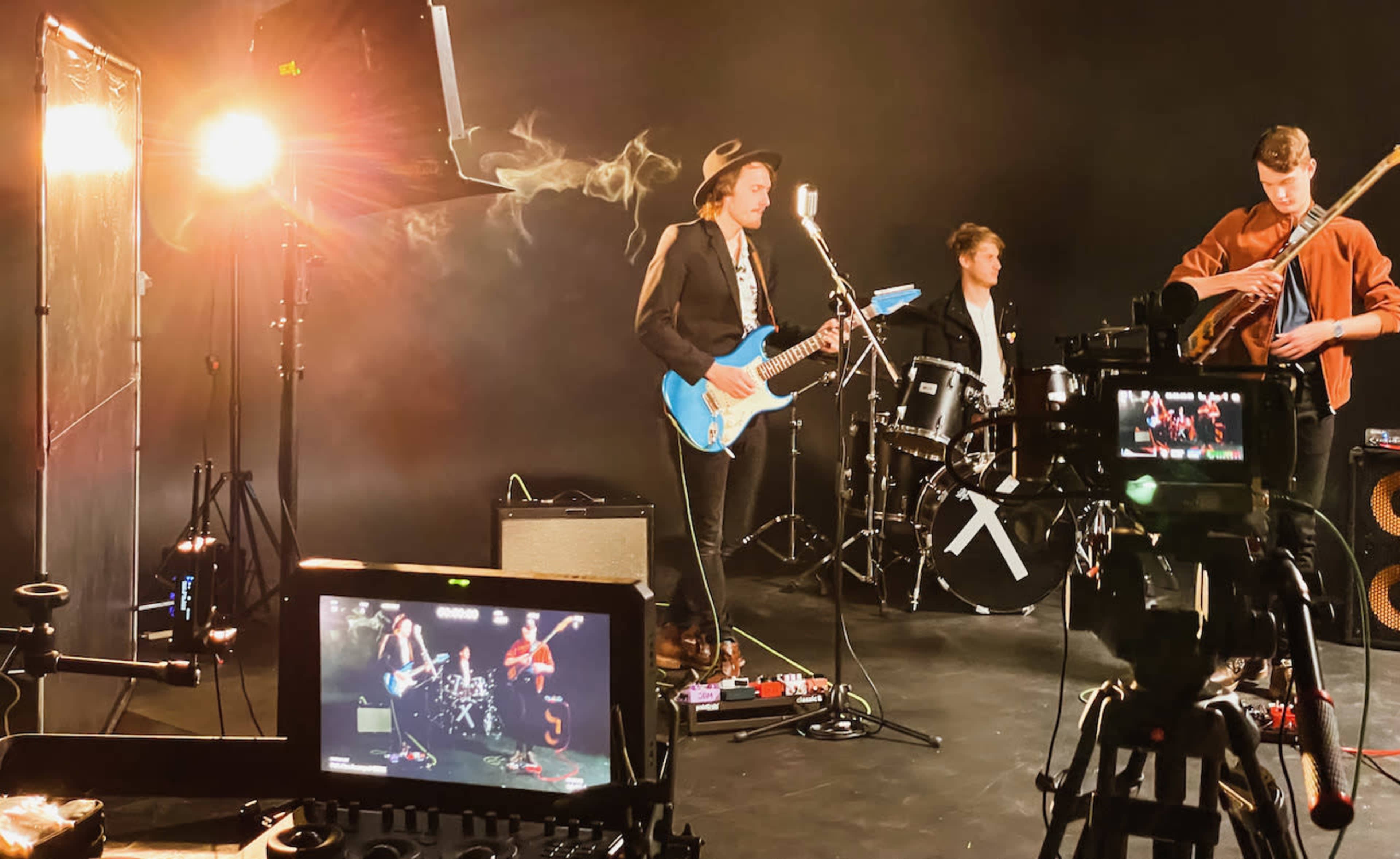A band performs on stage with a blue guitar, drums, and other instruments while cameras and lights set the scene.