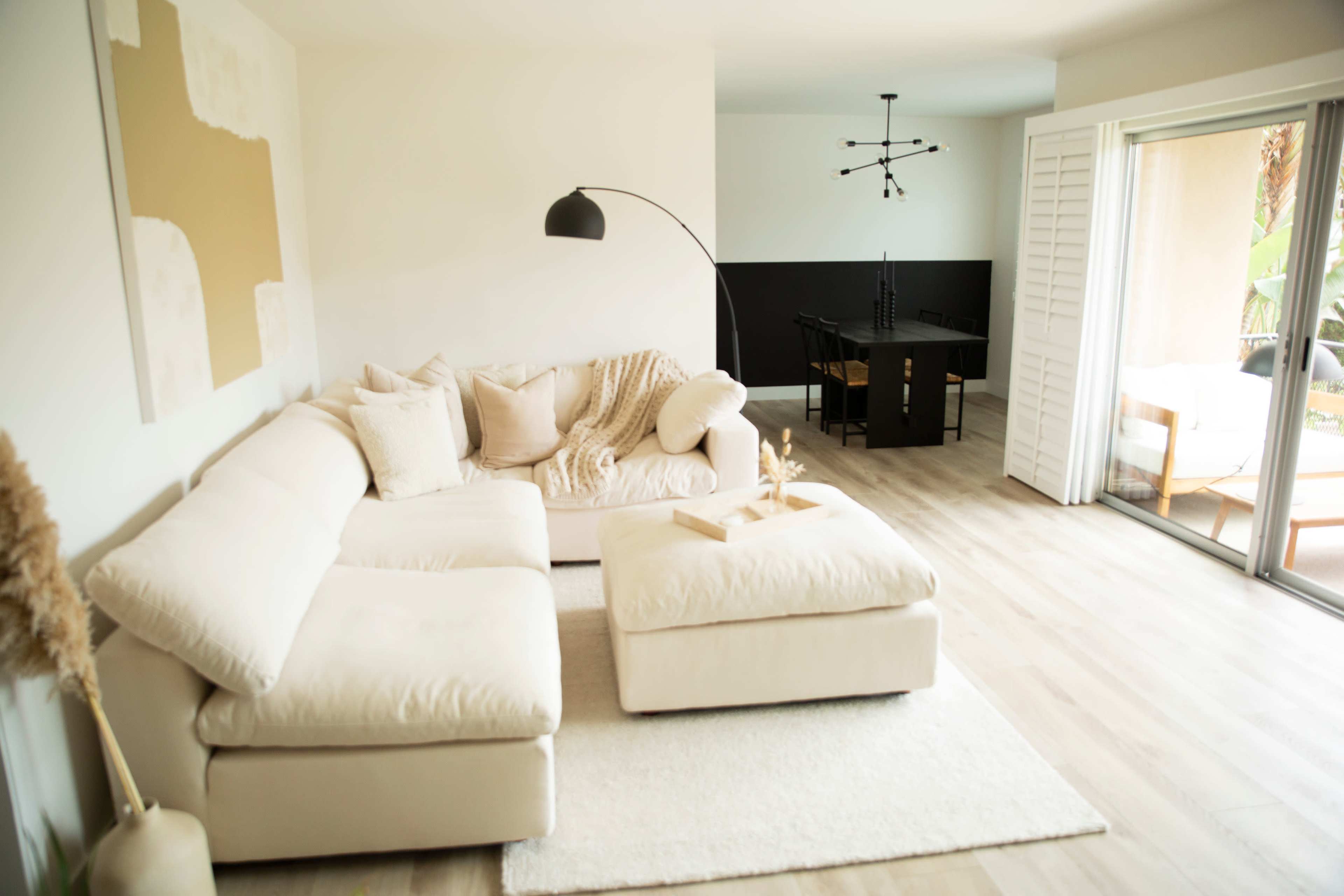 A light-colored living room featuring a large sectional sofa, an ottoman, and a dining area in the background with a modern chandelier.