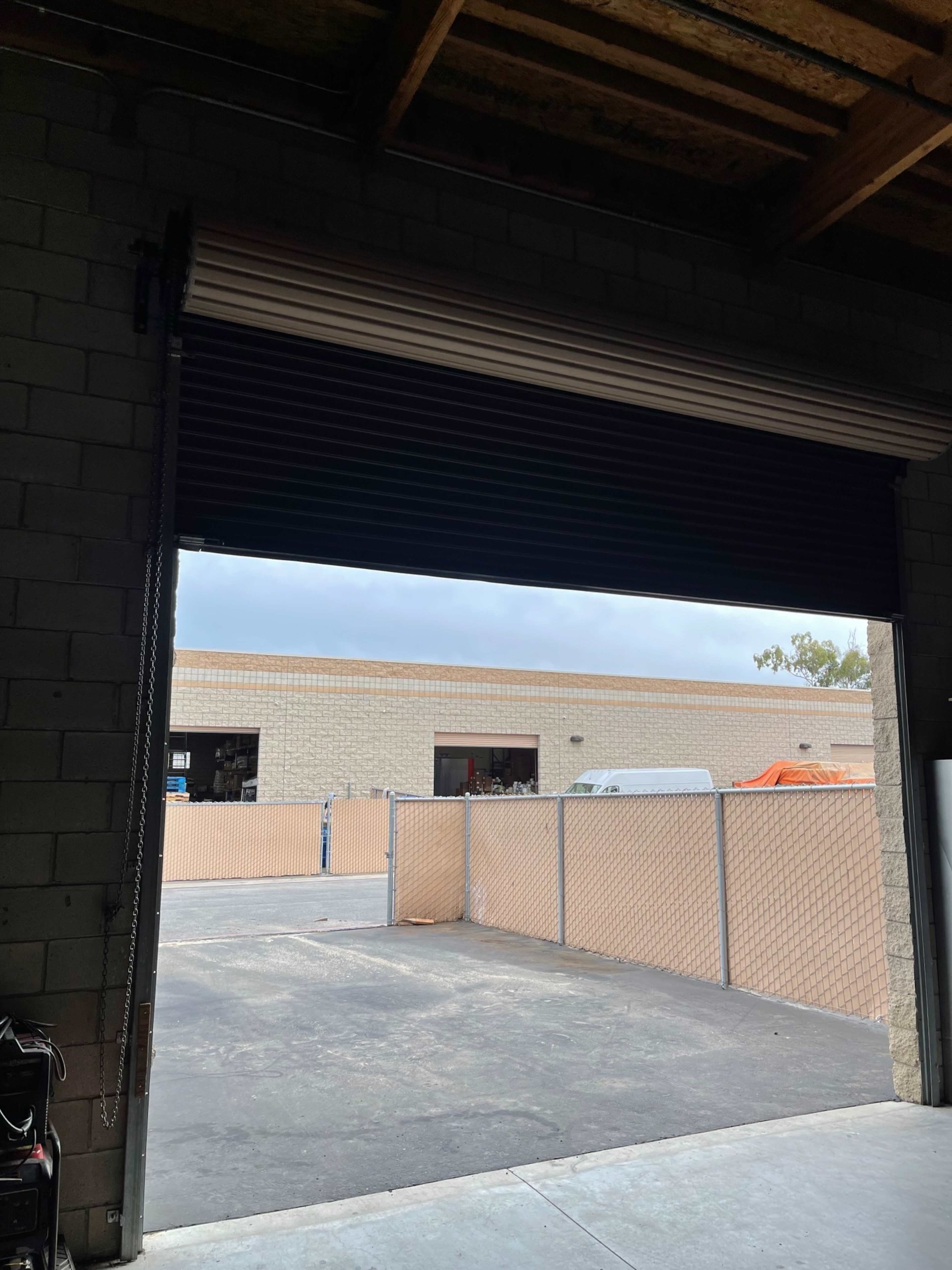 The interior of a garage or warehouse with a partially rolled-up metal door and a view of a fenced outdoor area beyond.