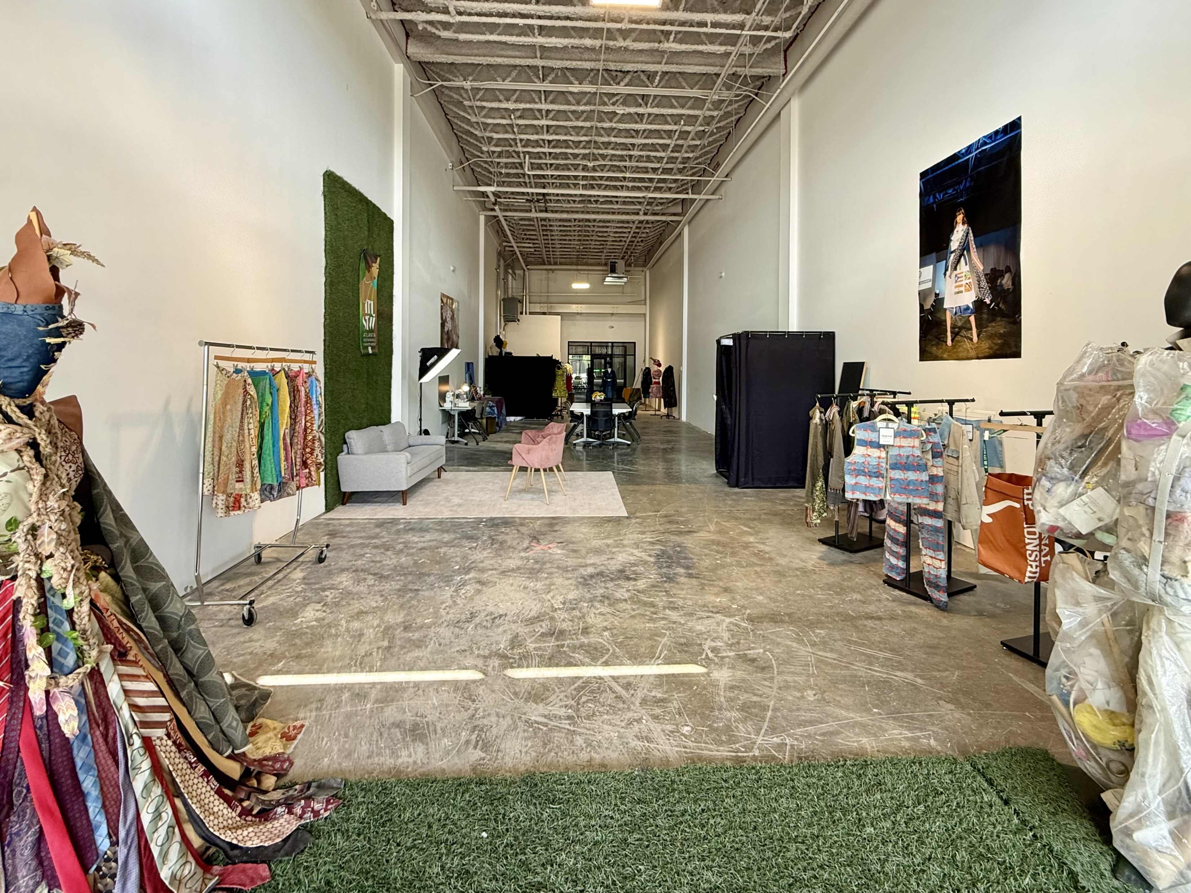 The image depicts a spacious, industrial-style interior with clothing racks on one side, greenery accents, and various furniture and equipment arranged throughout the area.