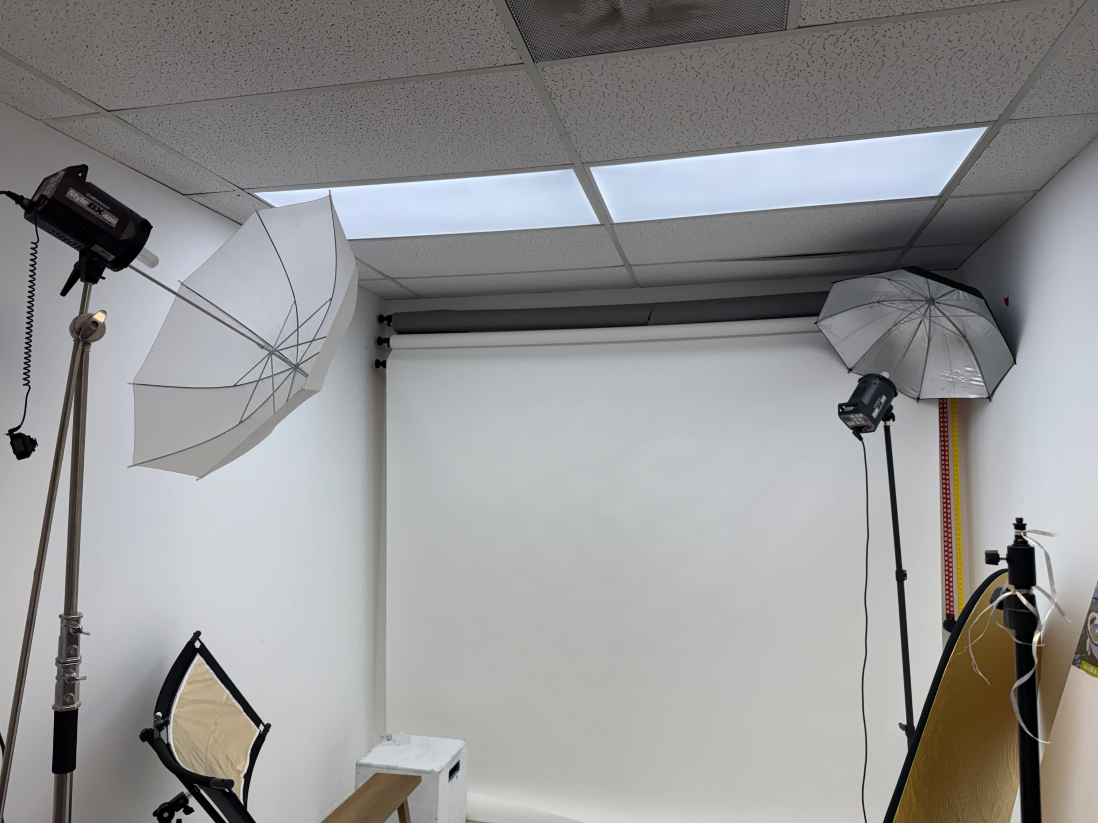 The image shows a photography studio with two light umbrellas, a camera tripod, and a plain white backdrop.
