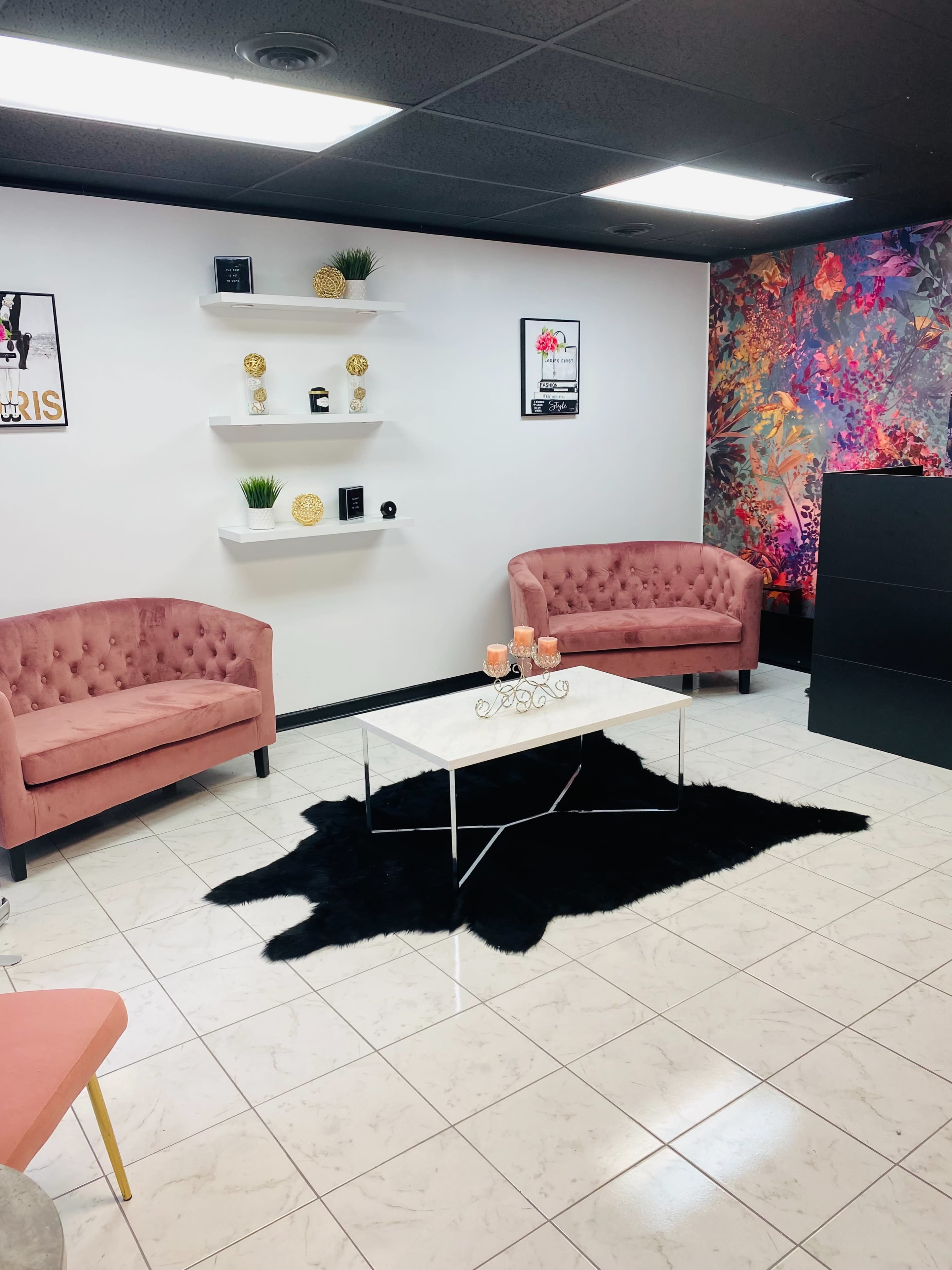 A modern waiting area with two pink tufted chairs, a white coffee table on a black rug, and decorative shelves on a white wall.