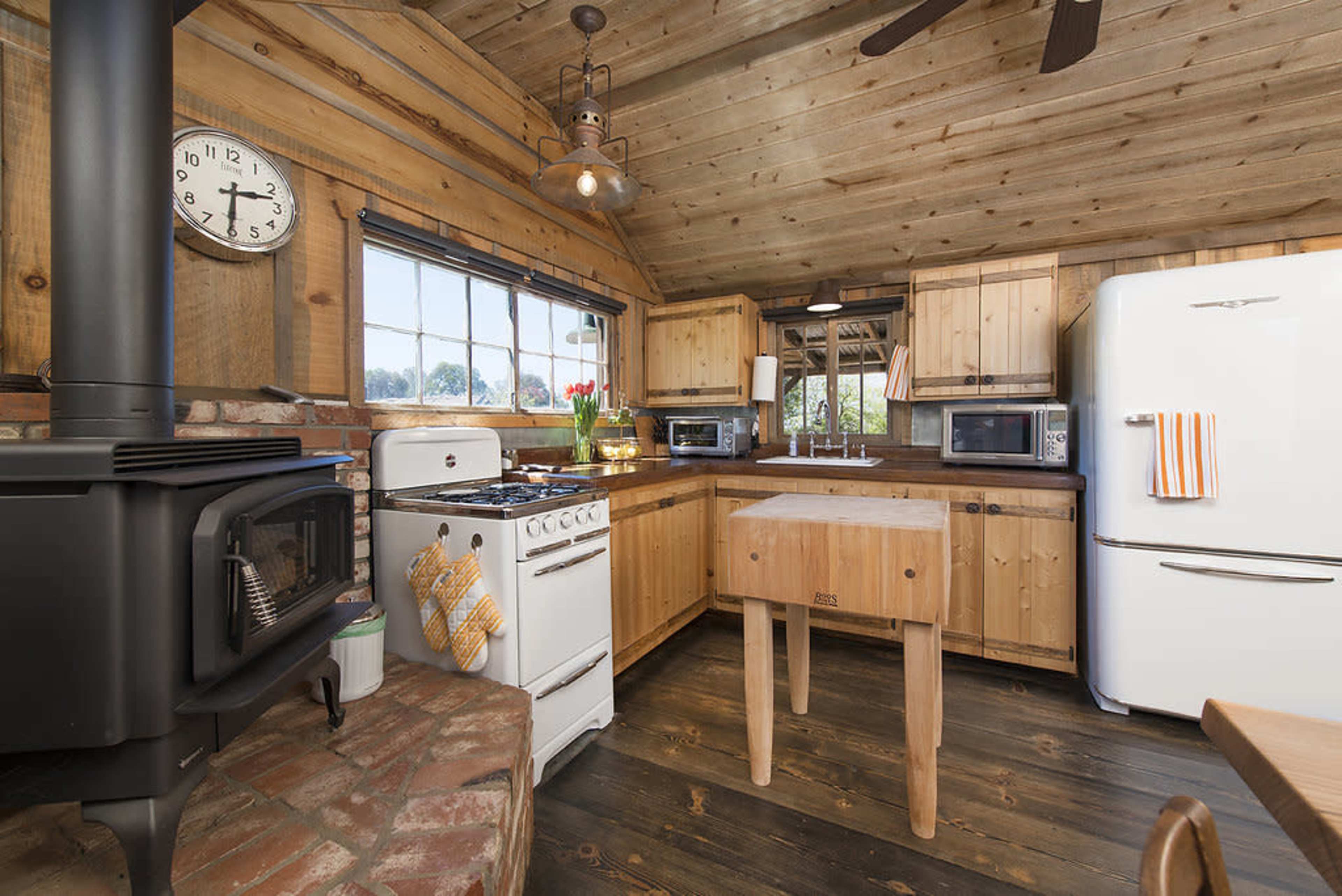 The kitchen features wooden walls, a stove, a refrigerator, and a central wooden table, all arranged around a large window.