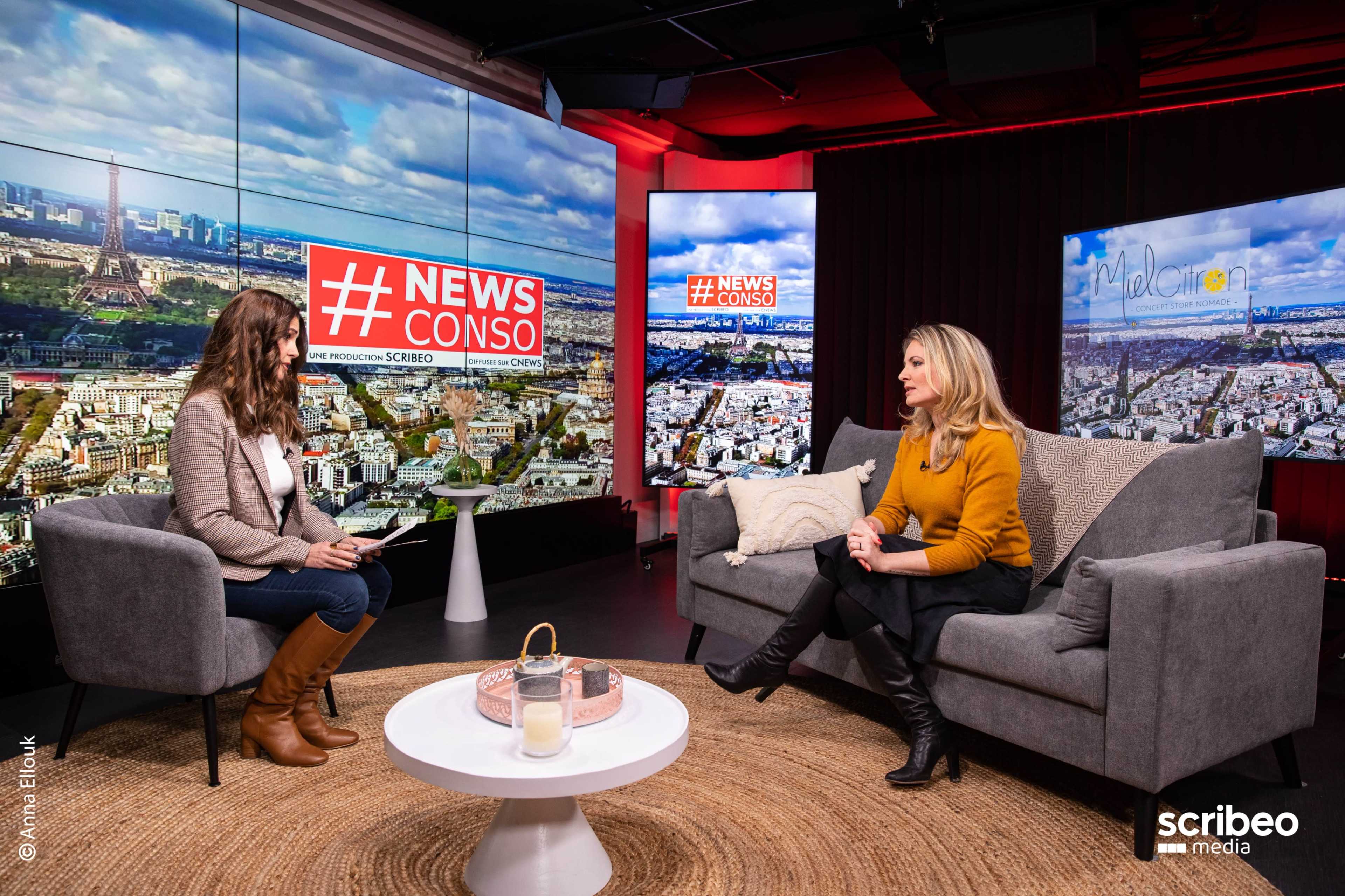 A host and guest are seated in a modern studio, discussing topics related to news, with a large screen displaying "#NEWS CONSO" in the background.