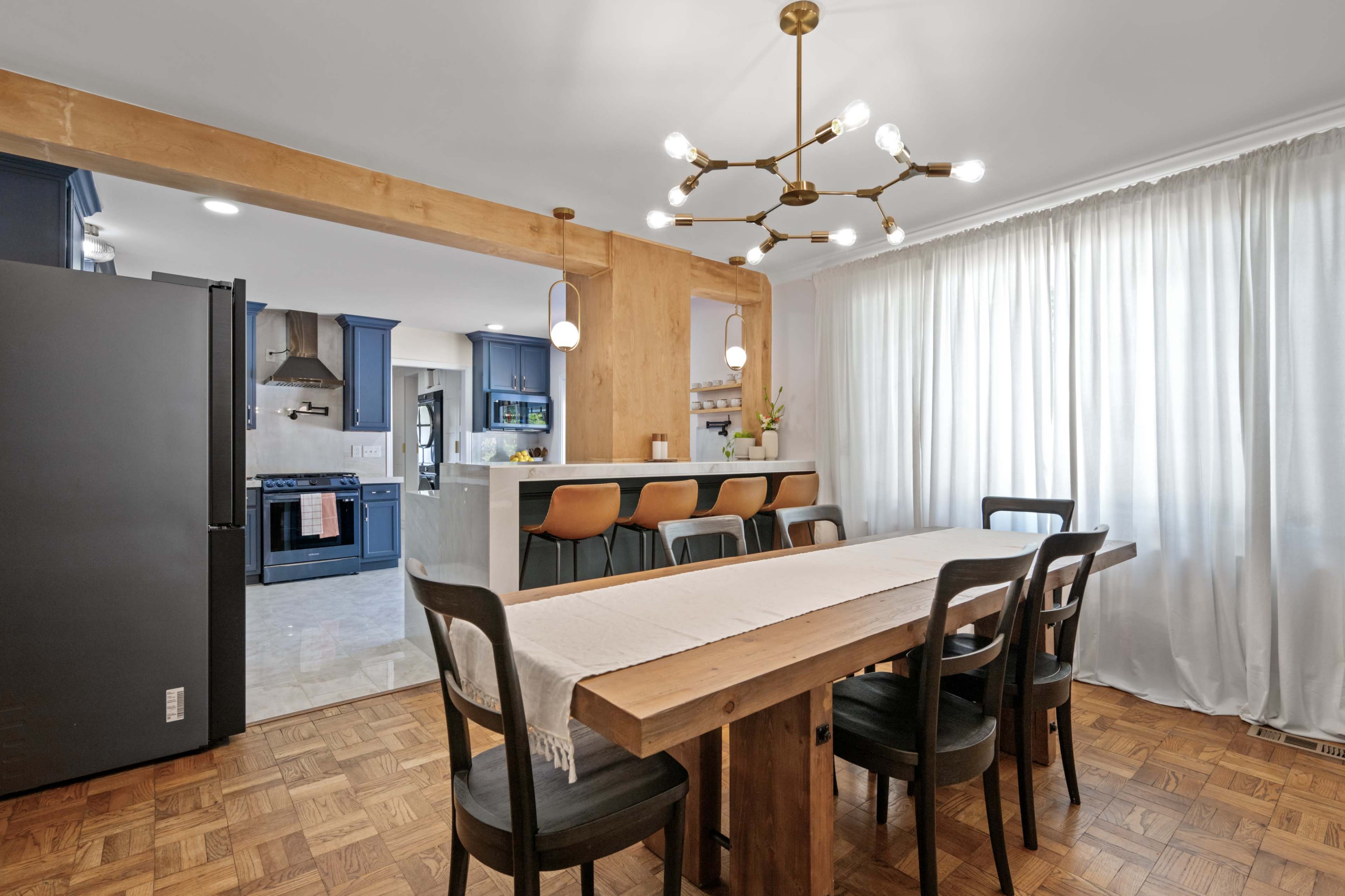 A modern kitchen and dining area featuring a wooden dining table, black chairs, blue cabinetry, and a well-lit space with a white curtain.