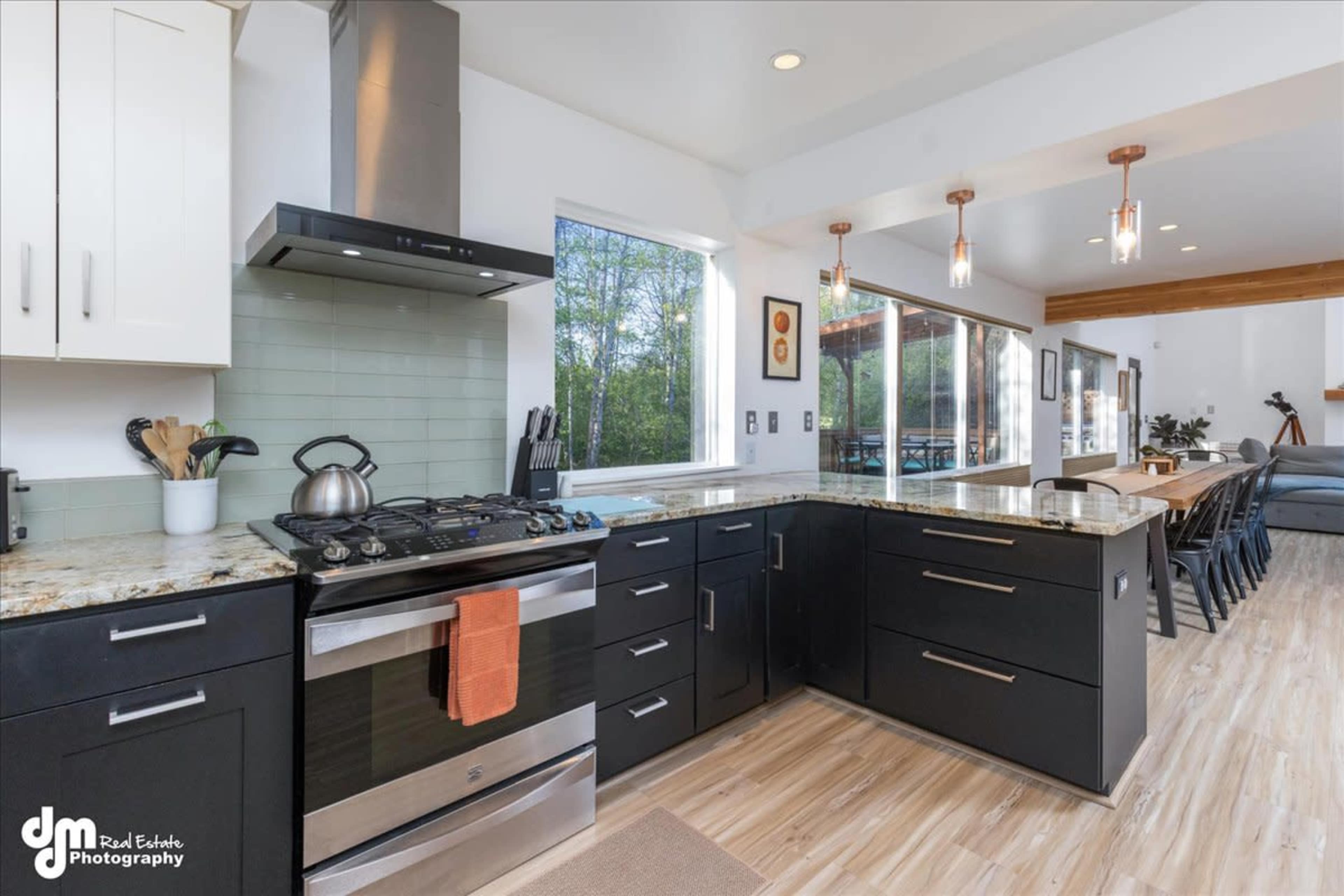 A modern kitchen with black cabinets, a stainless steel stove, and a granite countertop, adjacent to a dining area with pendant lighting.
