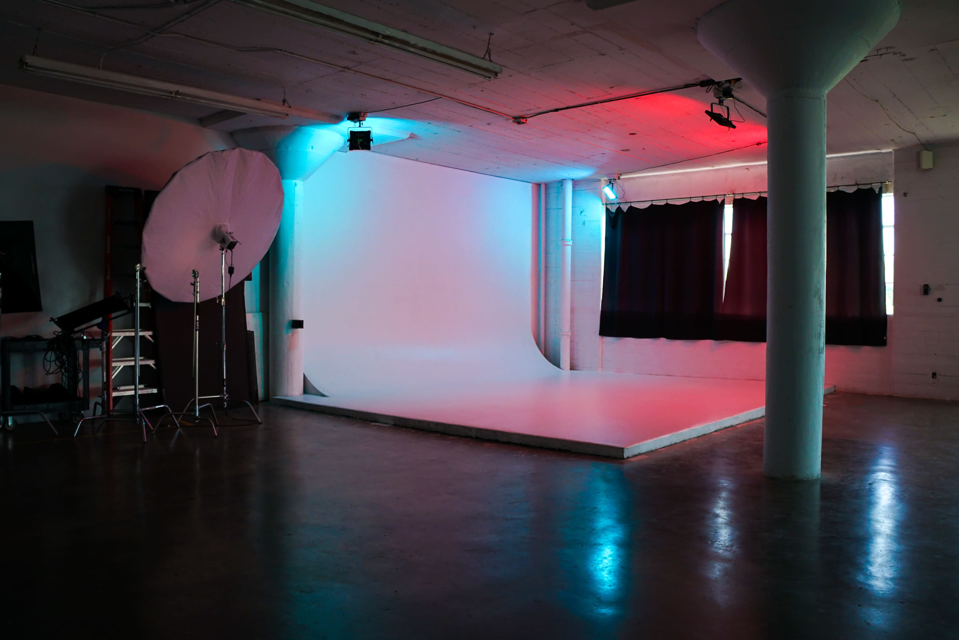 The image shows a photography studio with a lighted backdrop, a large umbrella softbox, and black curtains on windows.