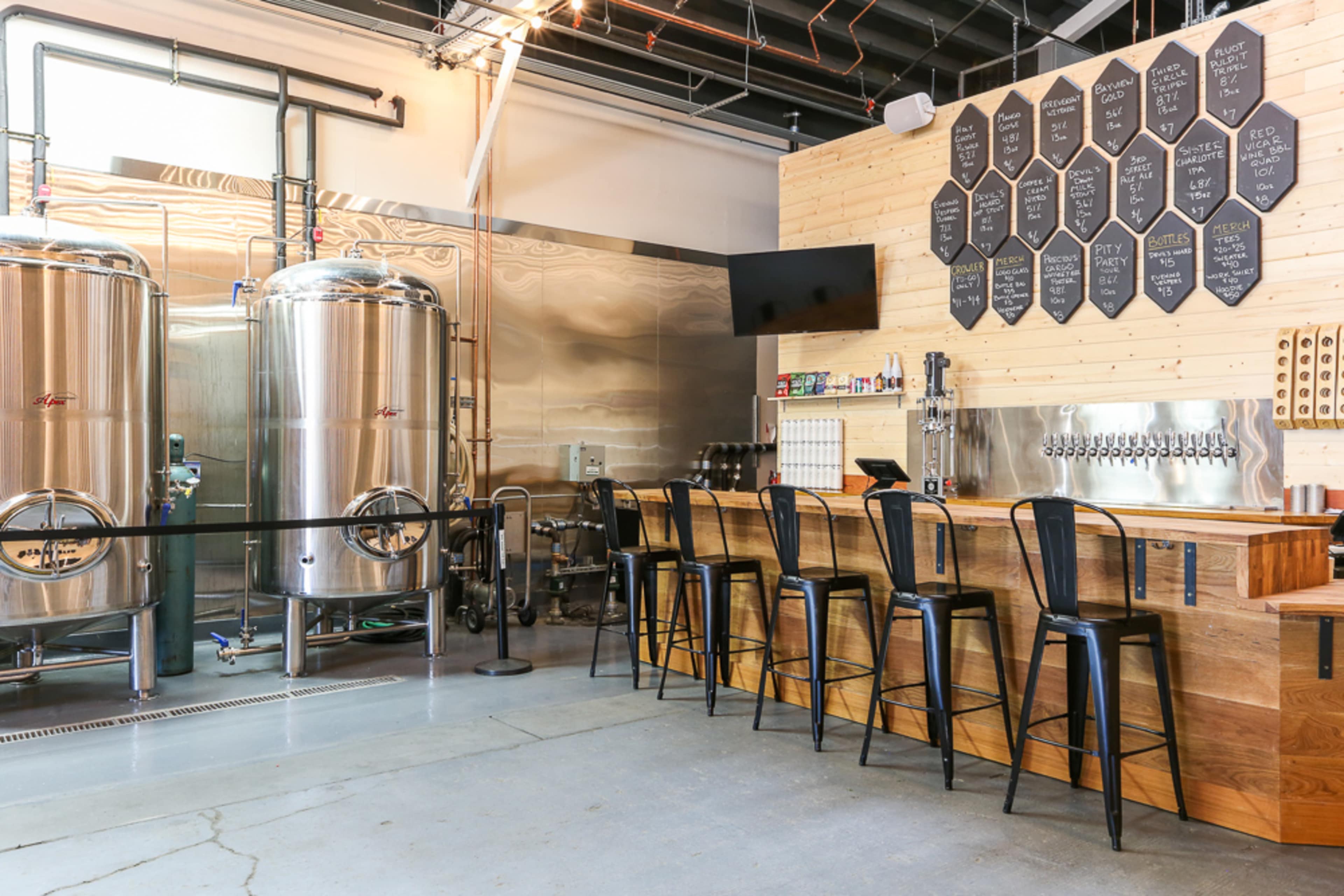 A brewery interior featuring large silver fermentation tanks and a wooden bar with high stools, against a backdrop of wall-mounted chalkboards displaying various beer options.