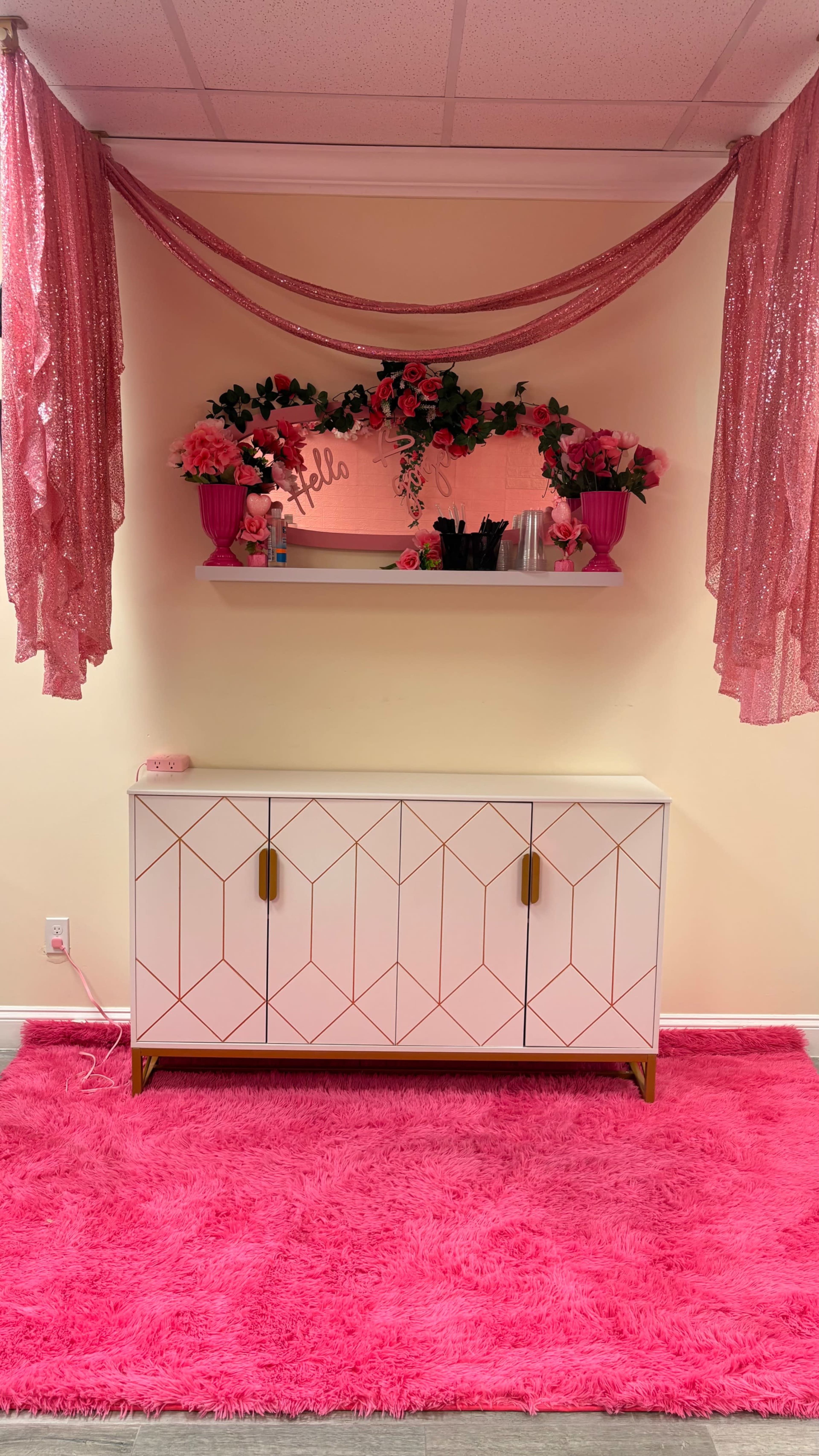The image shows a brightly decorated wall with pink drapes, a white sideboard with geometric patterns, and a pink shag rug underneath.