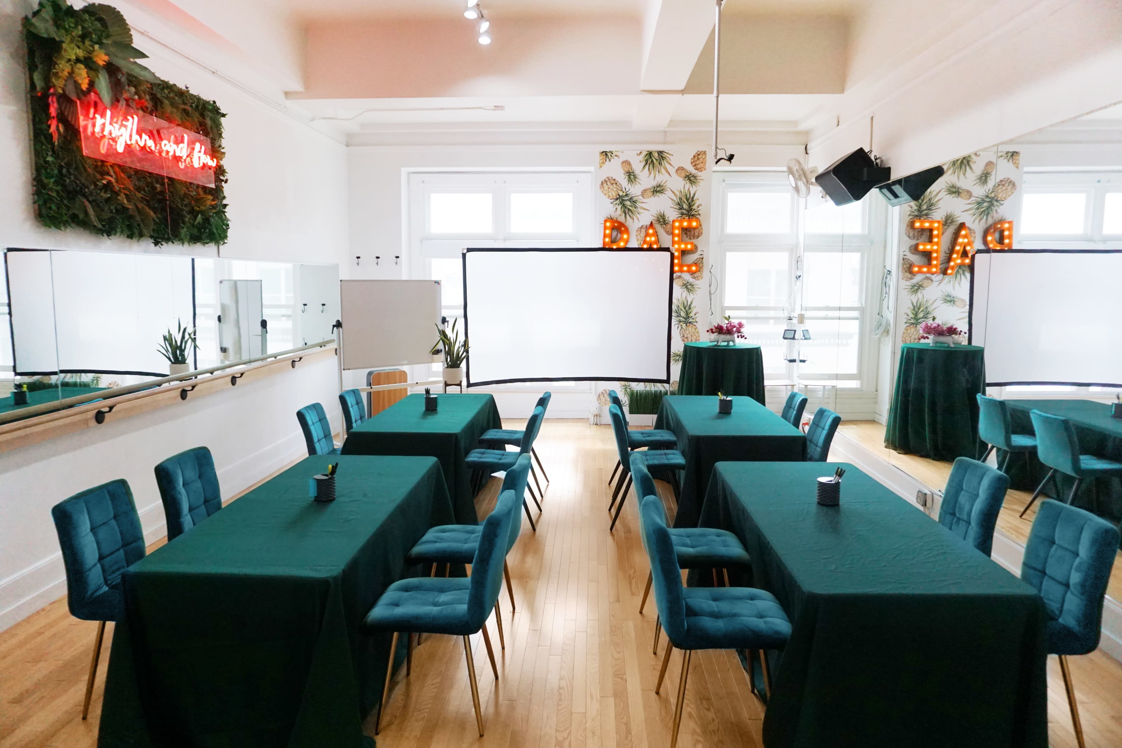 A room set up for a conference, featuring several tables covered with green cloths and chairs arranged around them, along with screens and decorative plants in the background.