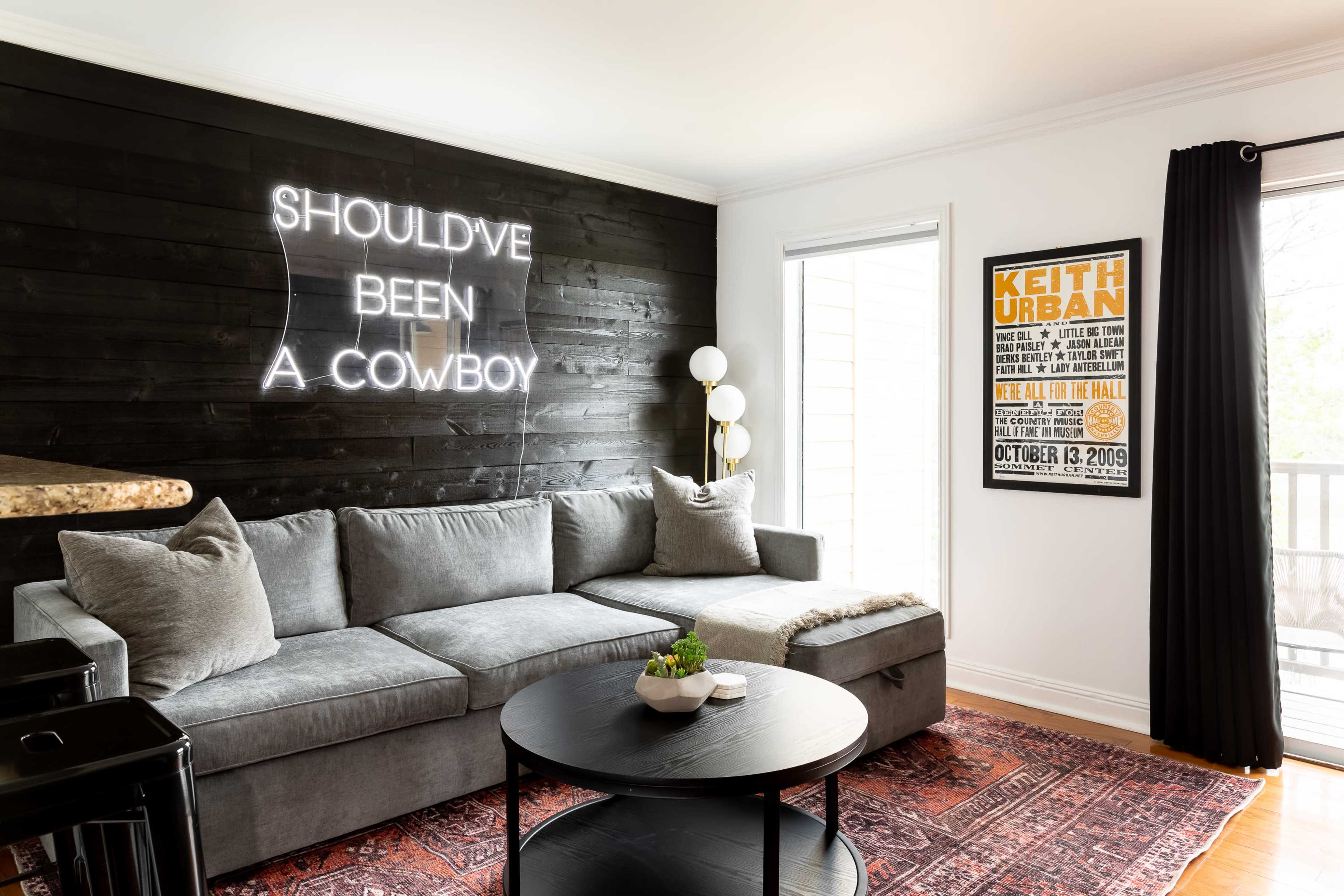 The living room features a gray sectional sofa, a round black coffee table, a neon sign on the wall that reads "SHOULD'VE BEEN A COWBOY," and a framed poster of a concert by Keith Urban.