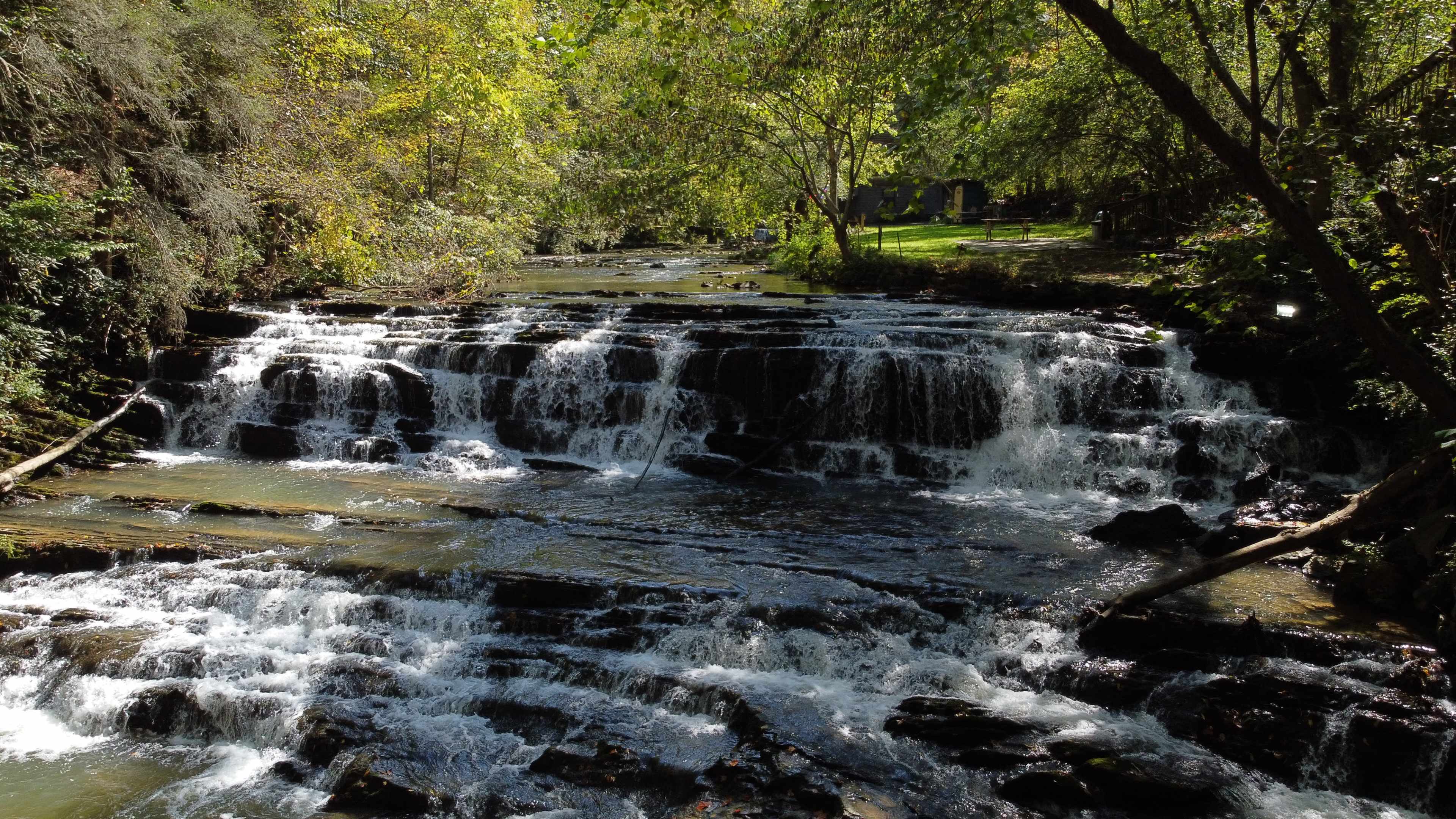 A waterfall cascades over rocky steps surrounded by lush greenery and trees.