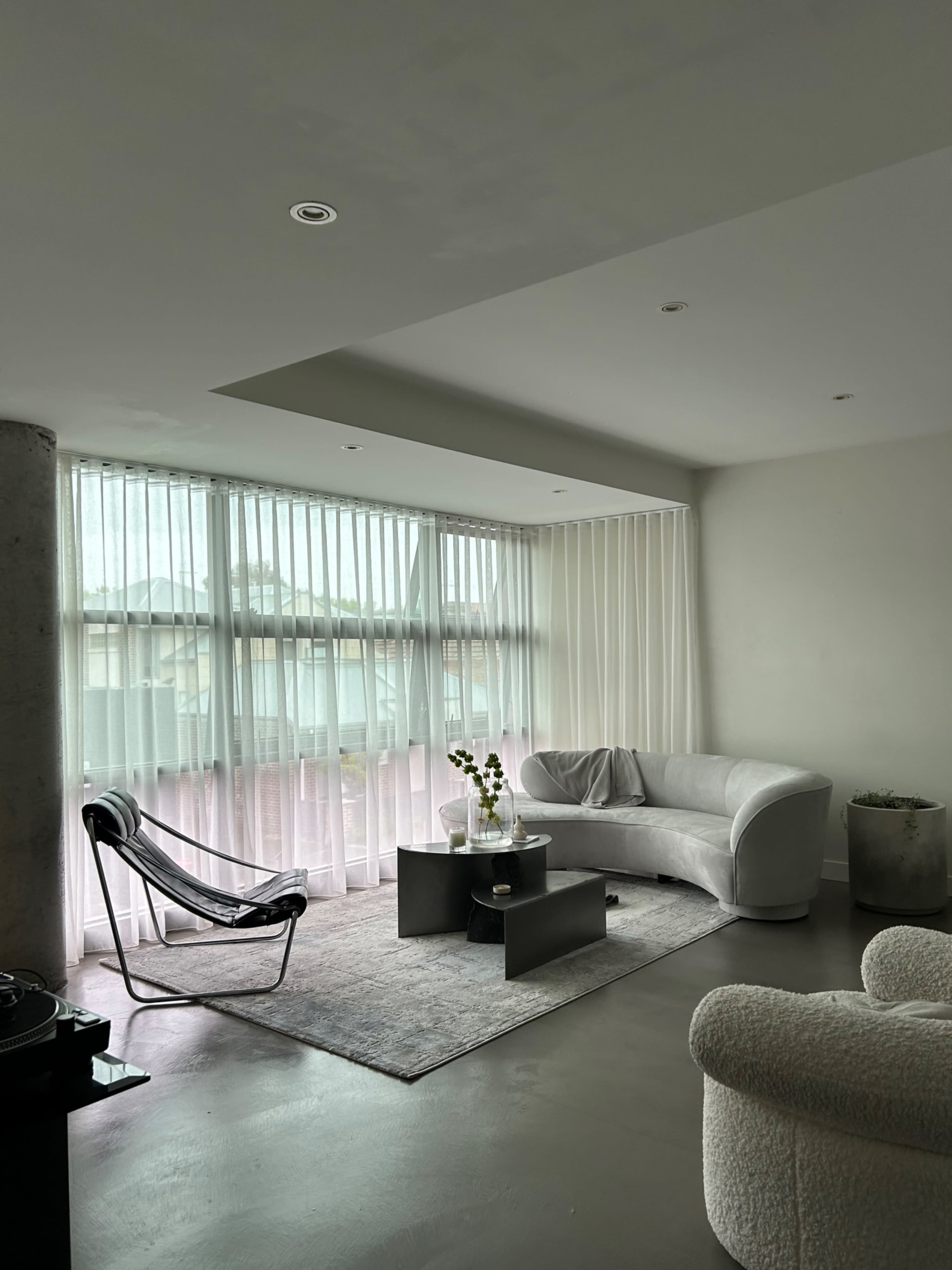 A modern living room with a light-colored couch, a black coffee table, and large windows covered by sheer white curtains.