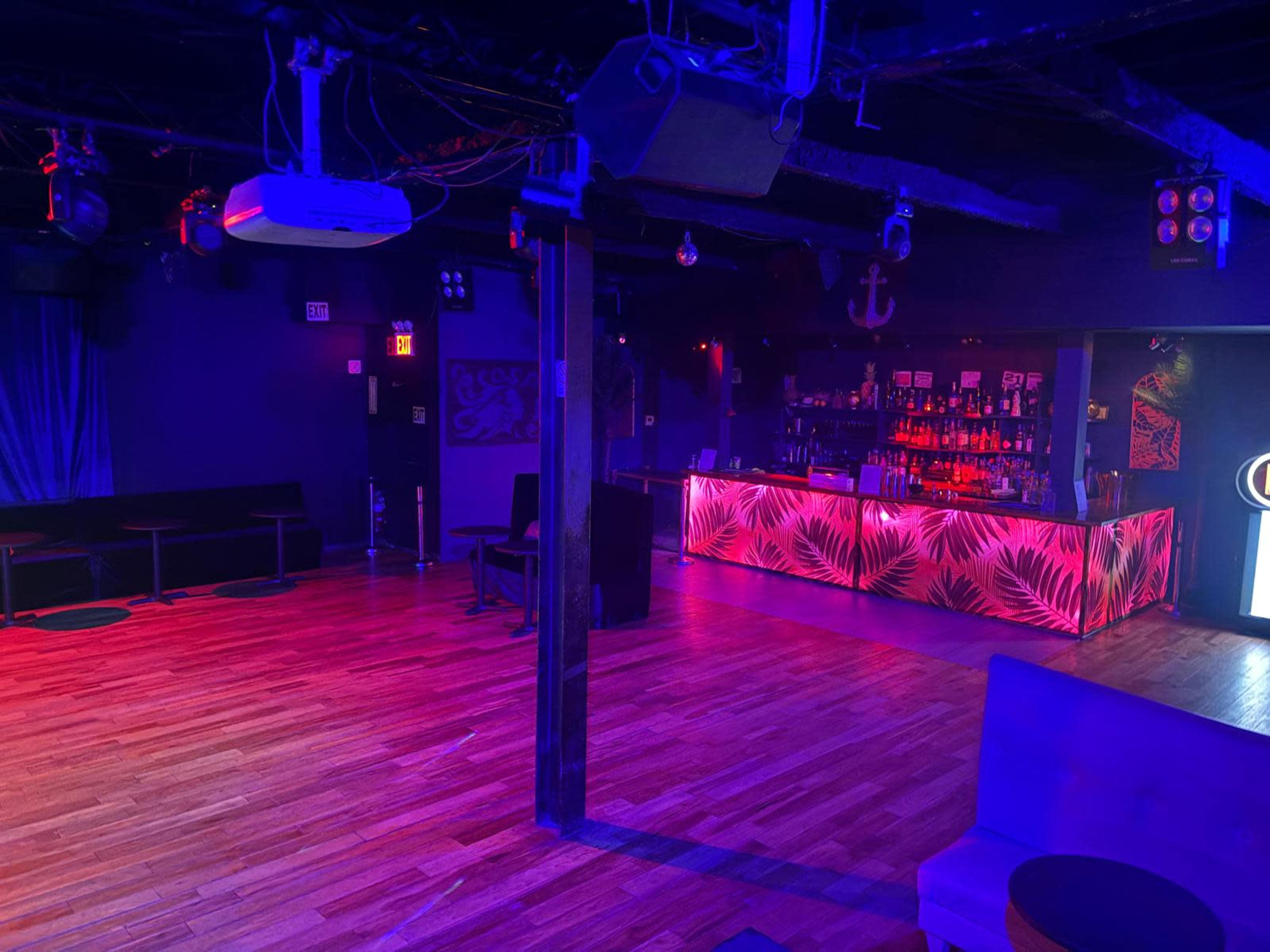 The image shows a dimly lit nightclub interior featuring a bar with colorful lighting and wooden flooring.