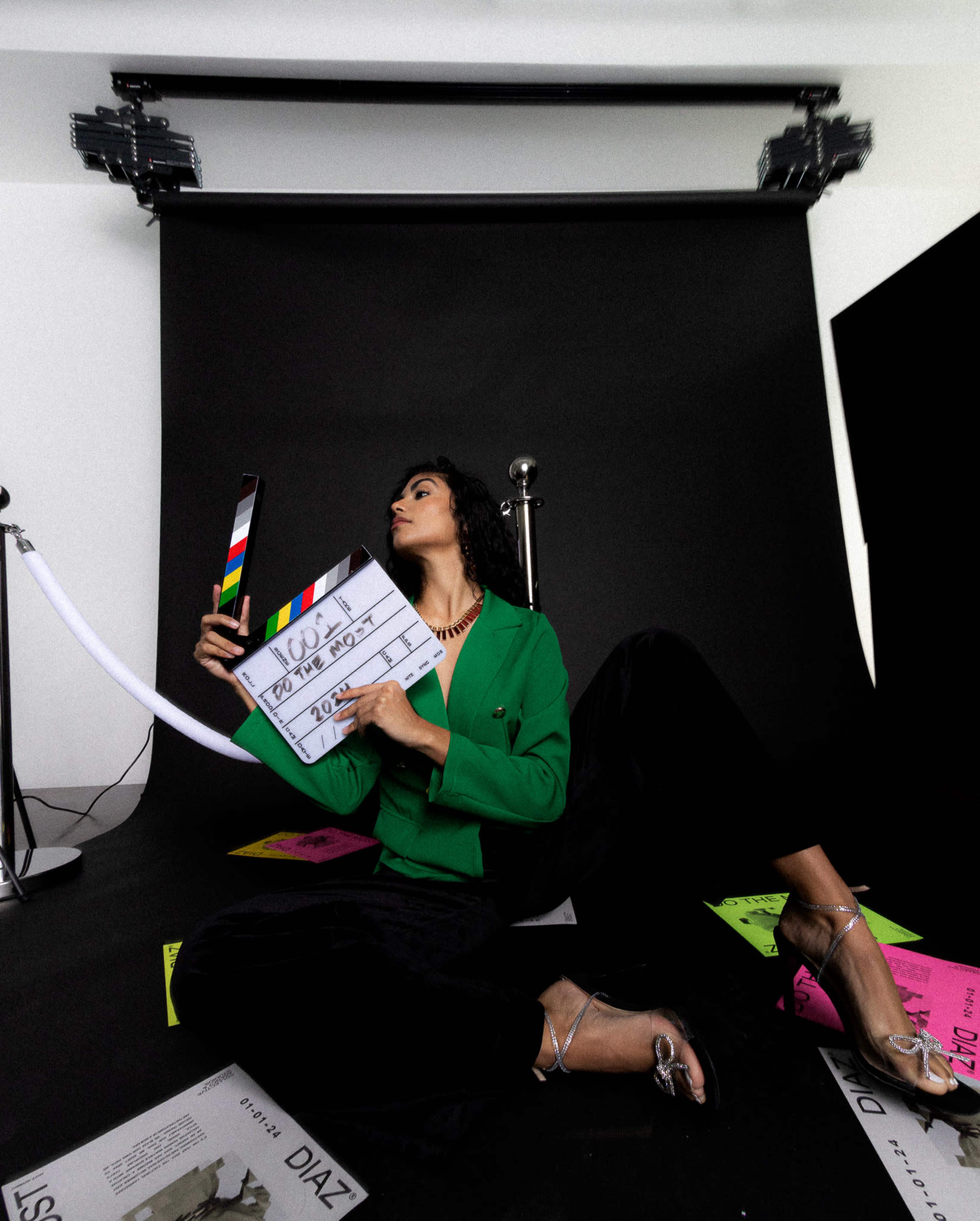A woman in a green blazer sits on the floor, holding a clapperboard amidst scattered magazines and colorful papers in front of a black backdrop.