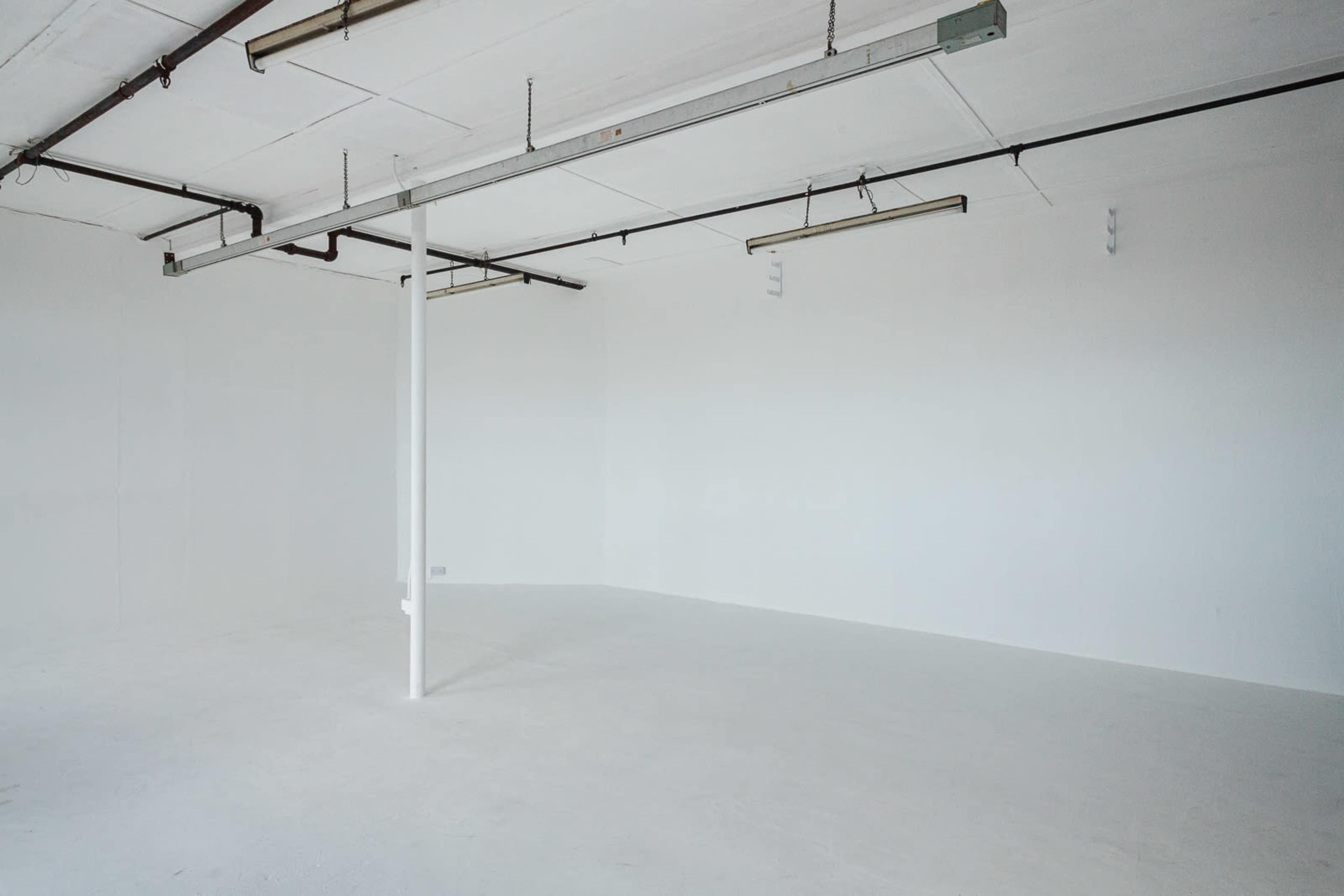 A spacious, empty room with white walls and flooring, featuring overhead lighting and exposed plumbing.