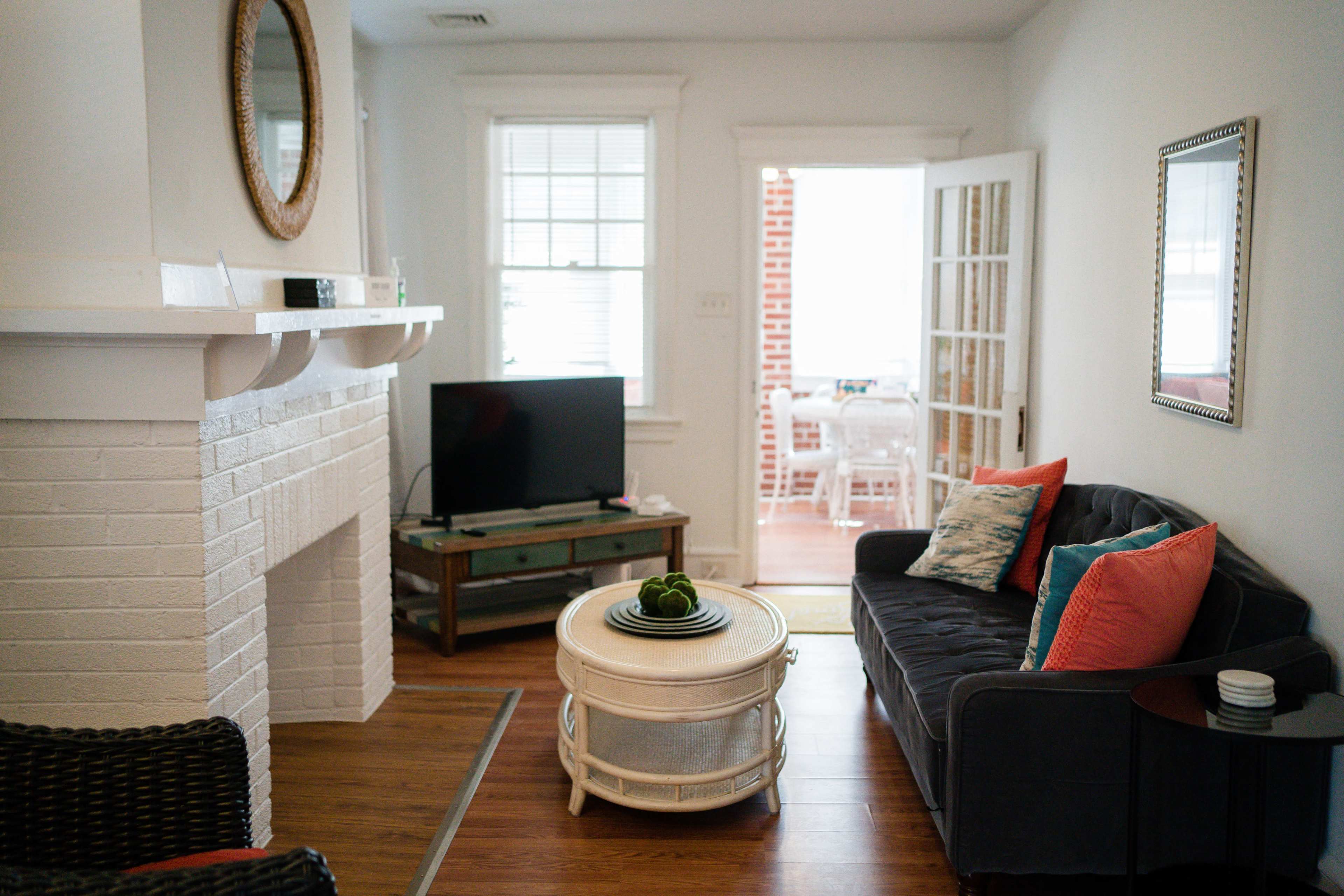 A living room with a gray couch, a coffee table, a television on a stand, and a view of a sunlit dining area through French doors.