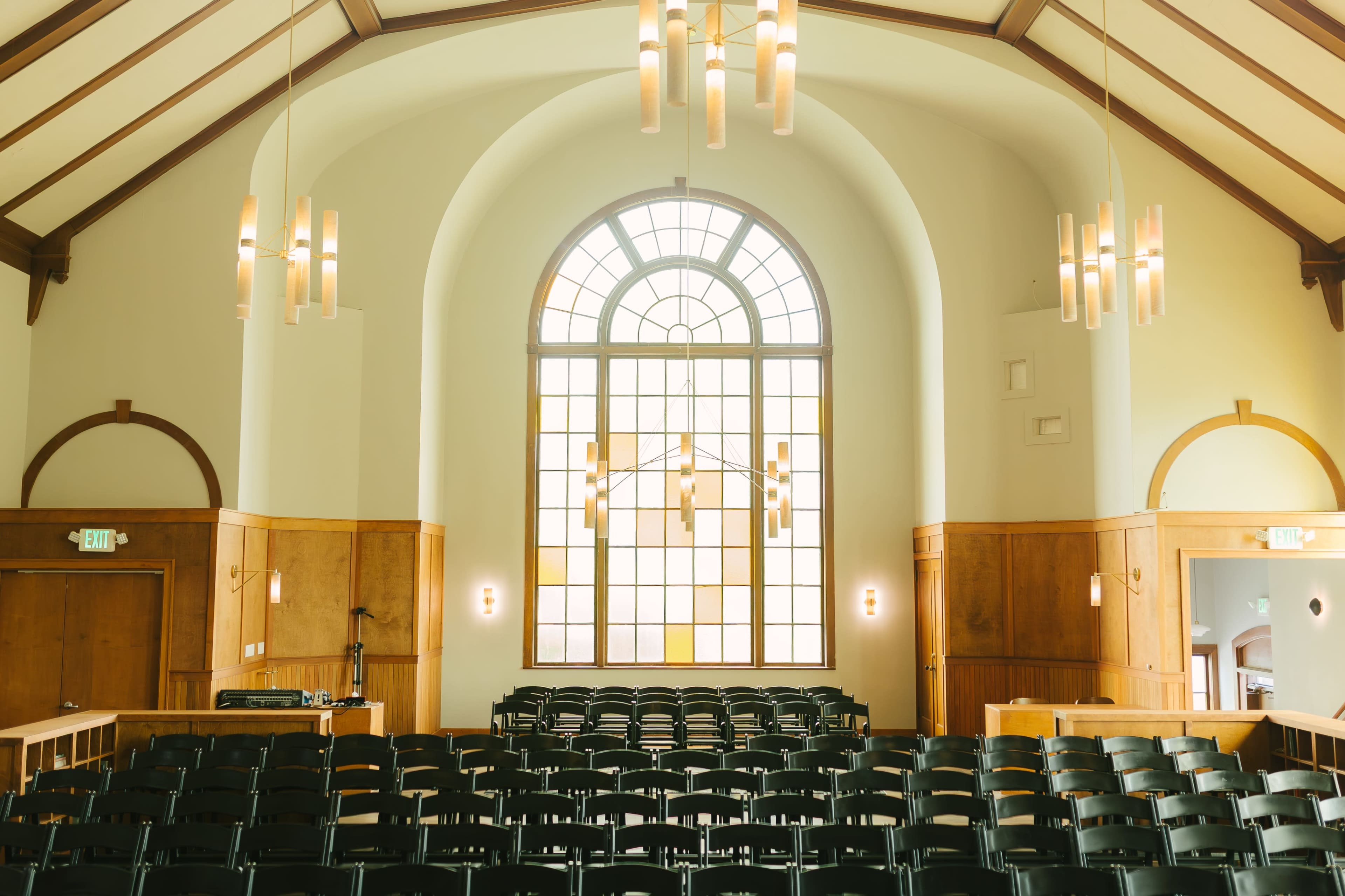 The image shows a spacious room with an arched window, wooden paneling, and rows of black chairs facing the front.