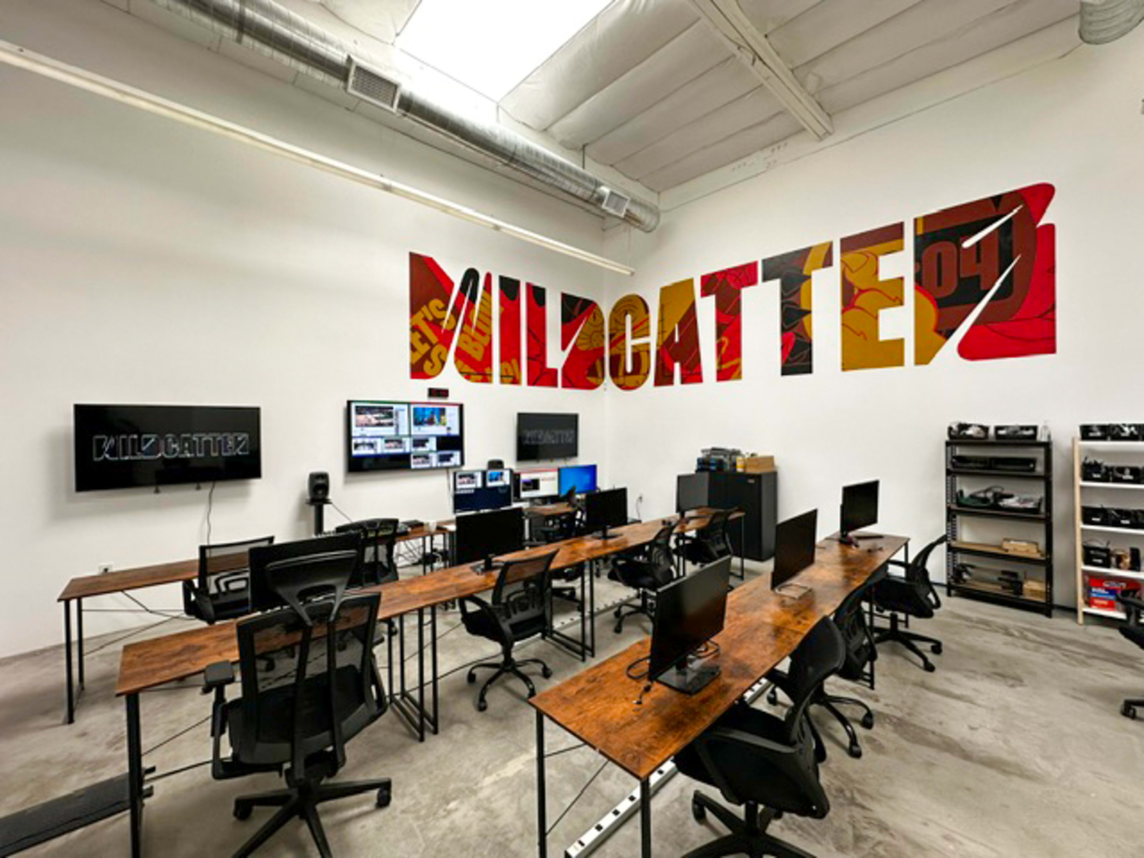 A modern office space featuring multiple workstations with computers, a large wall mural that says "WILDCATTEZ," and shelving units filled with equipment.