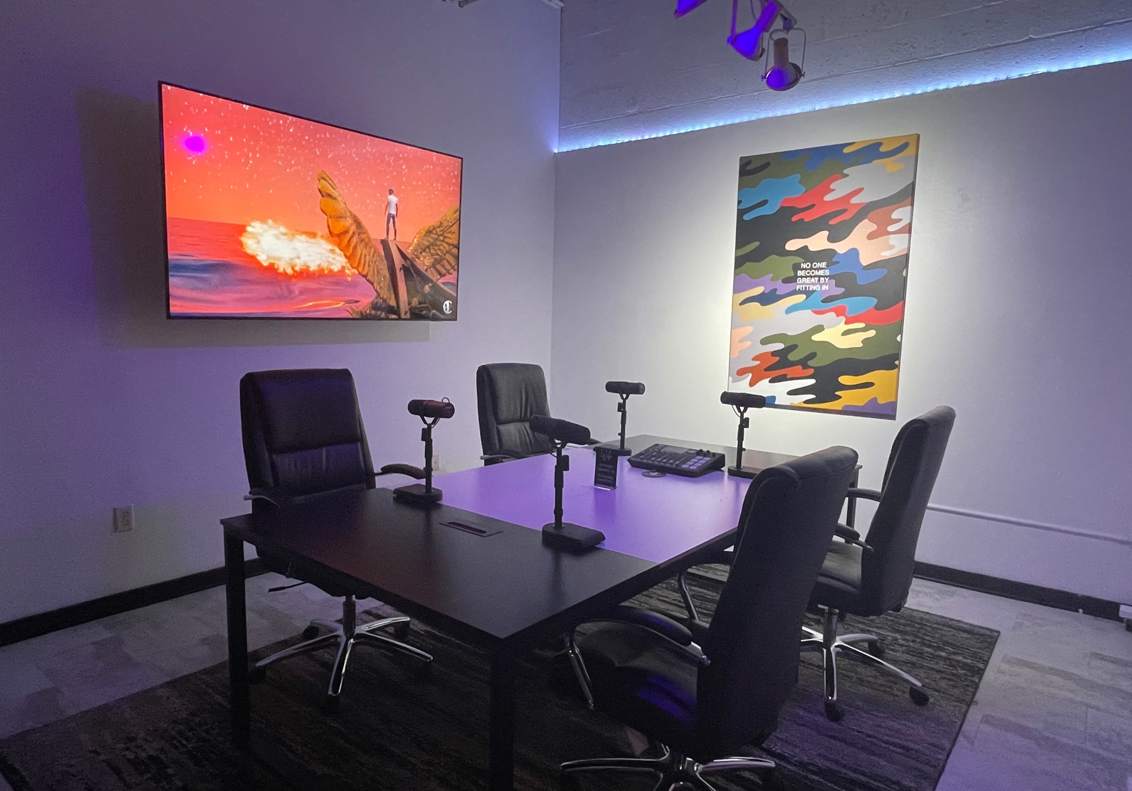 The image shows a modern office space with a large screen displaying colorful artwork, a wall adorned with a patterned piece, and a table surrounded by four black chairs and microphones.